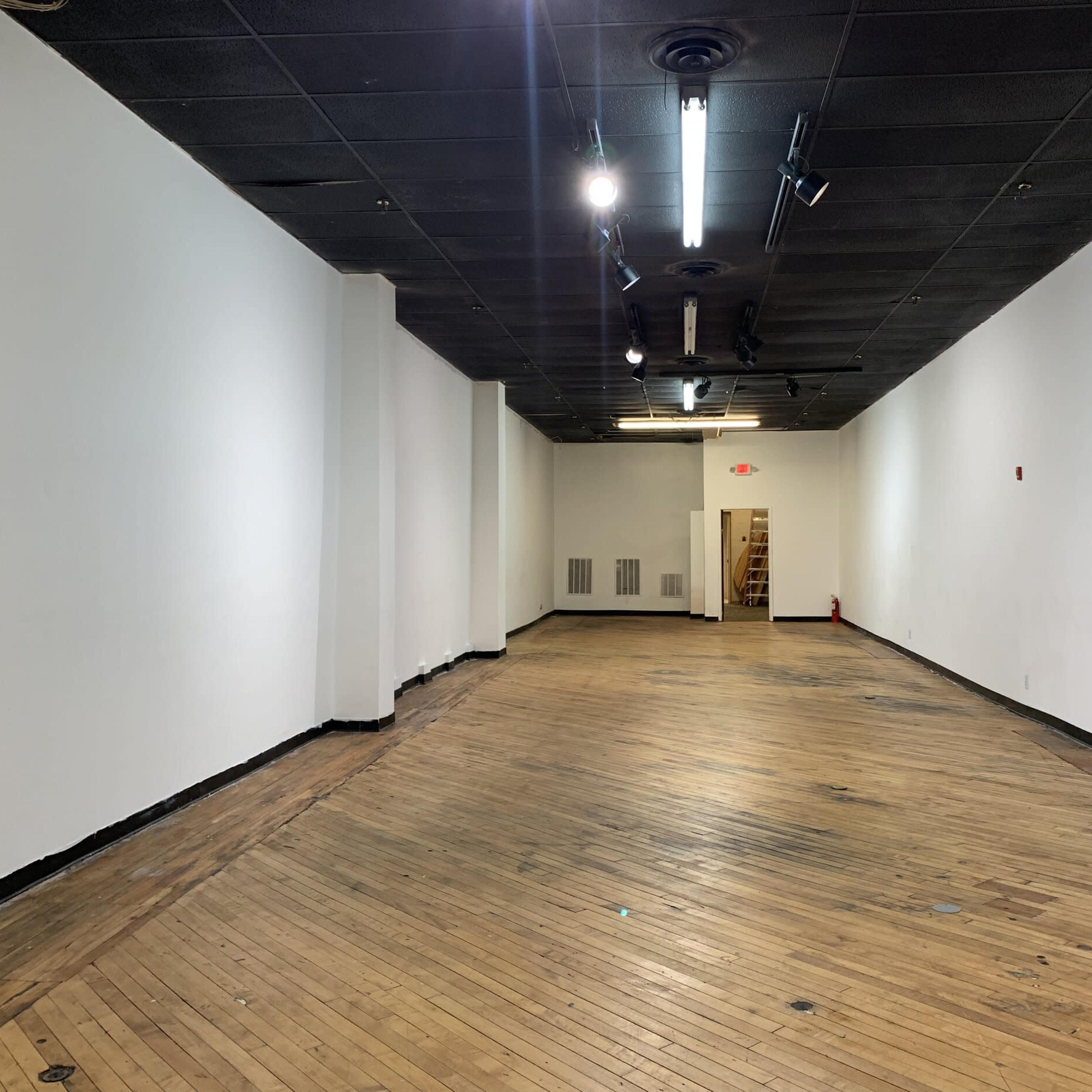 A large, empty interior space with wooden floors and plain white walls, featuring track lighting on the ceiling.