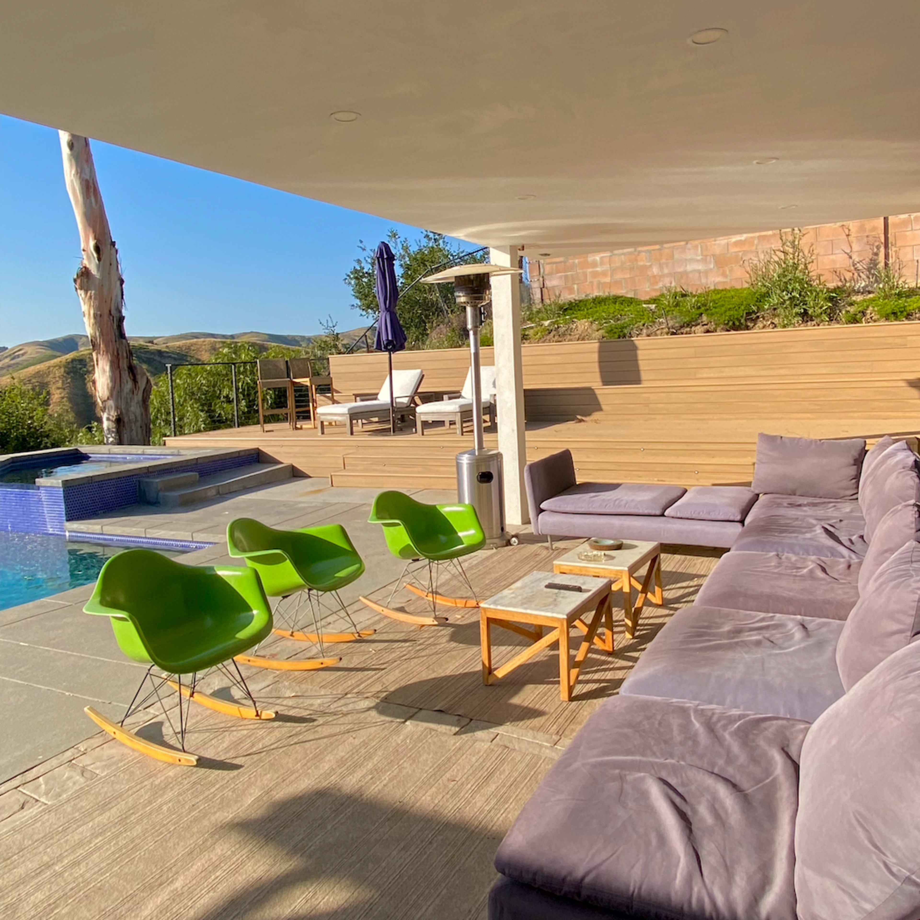 A modern outdoor lounge area features a large sectional sofa, four green chairs, a small table, and a pool in the background.