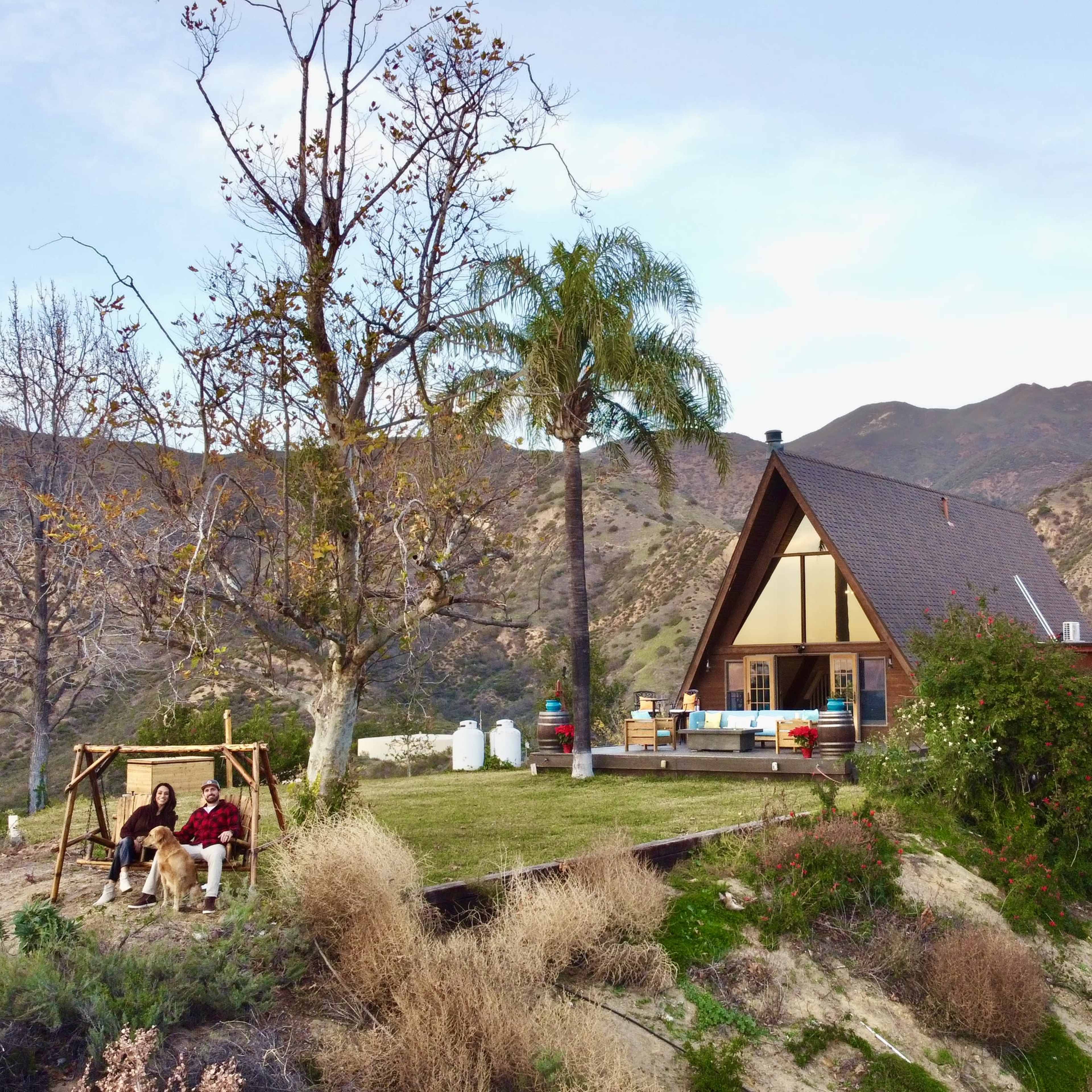 A wooden house with a triangular roof sits on a hill, surrounded by trees and mountains, with a couple sitting on a swing nearby.
