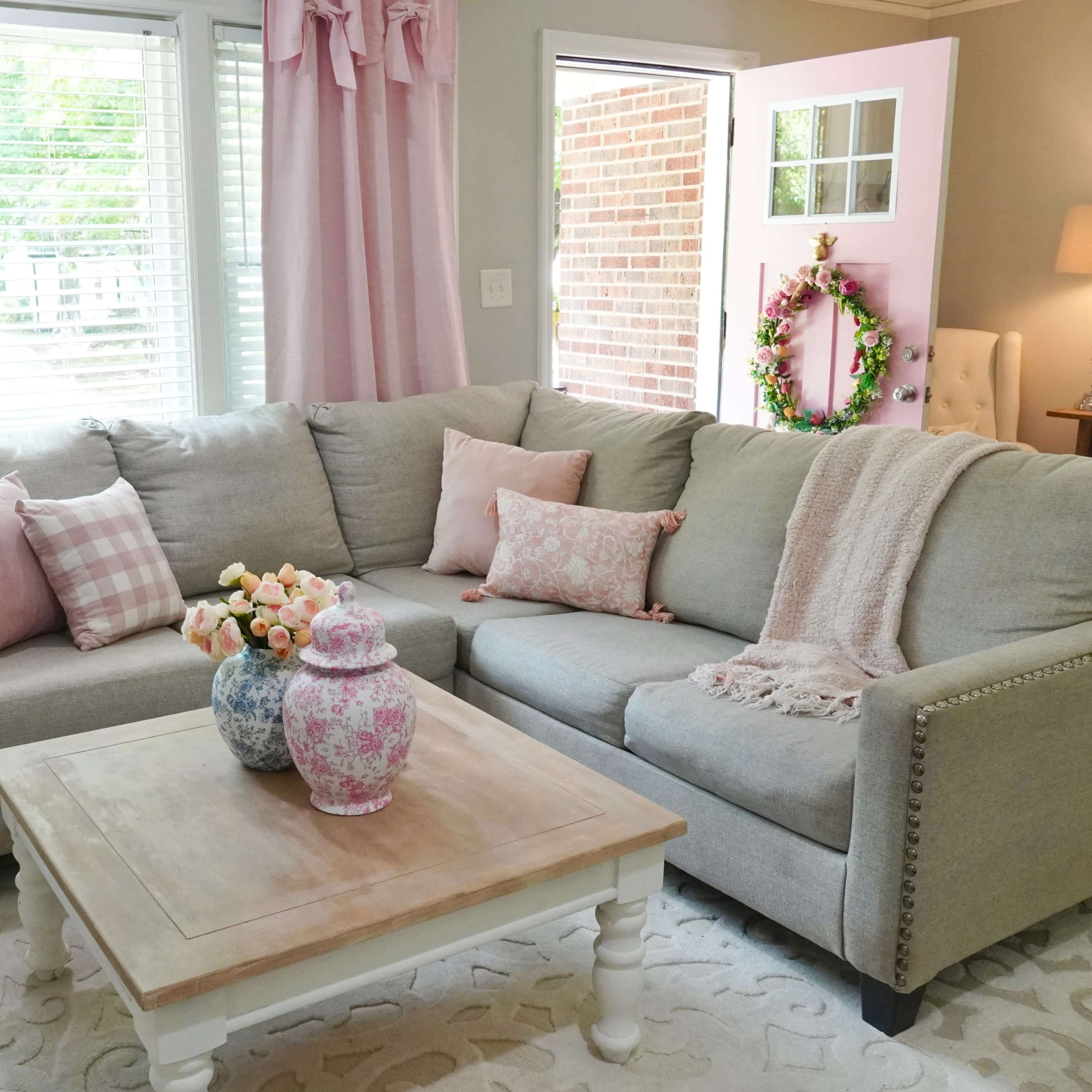 A cozy living room featuring a gray sectional sofa with decorative pillows, a wooden coffee table with floral vases, and large windows allowing natural light.
