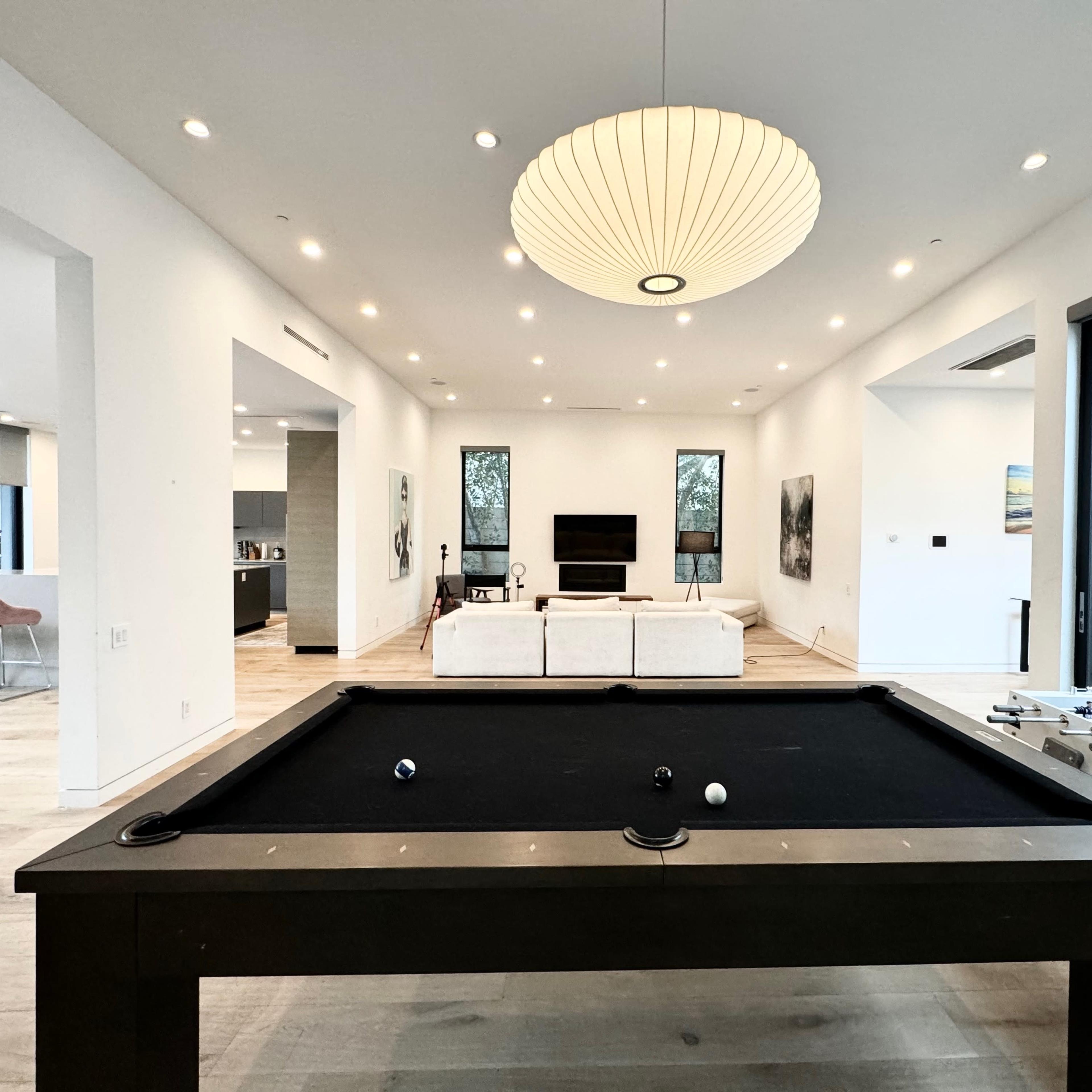 A billiard table is positioned in the foreground of a spacious room that features a living area with a sofa and a television in the background.