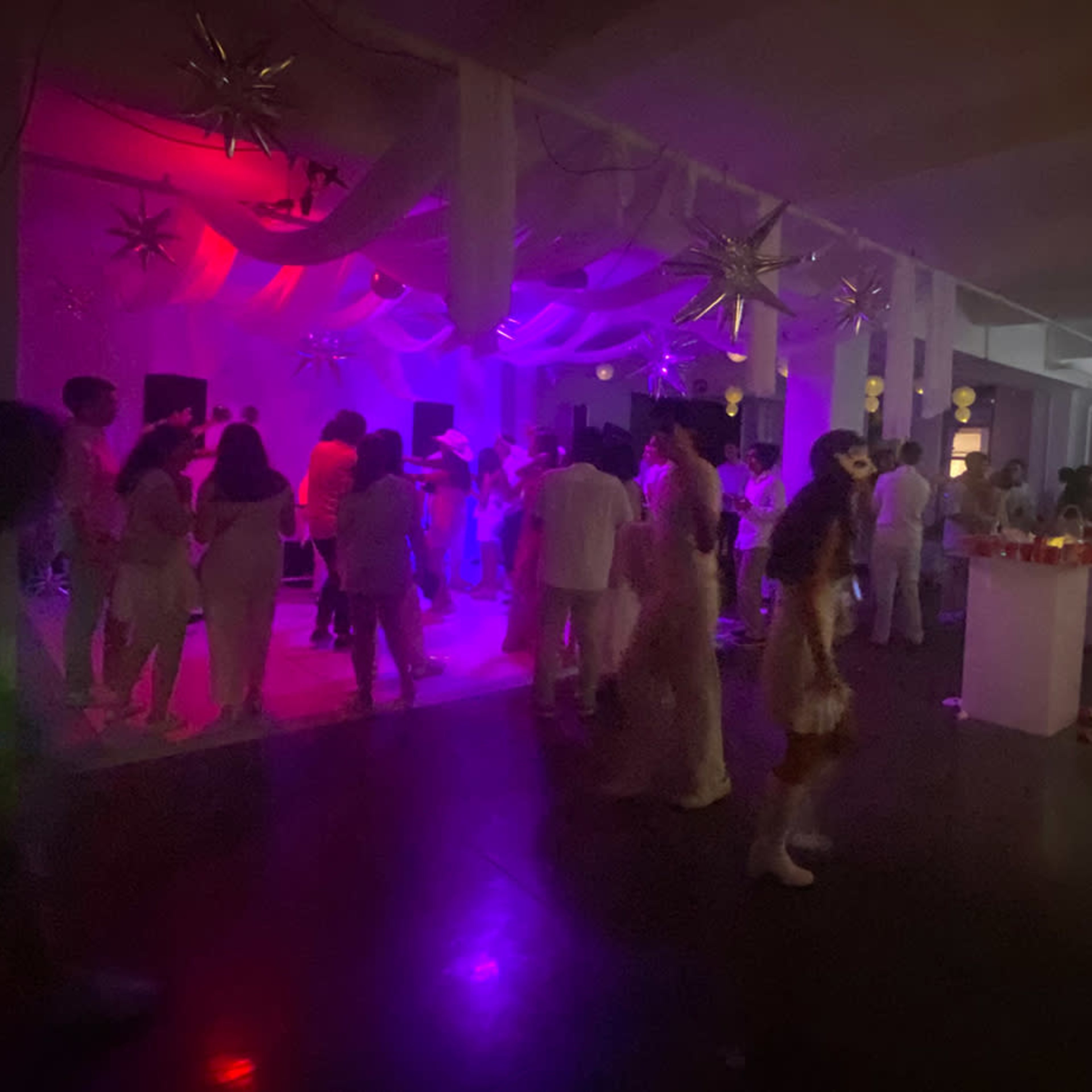 A lively indoor party gathers guests dressed in white, illuminated by colorful lights and decorations while music plays in the background.