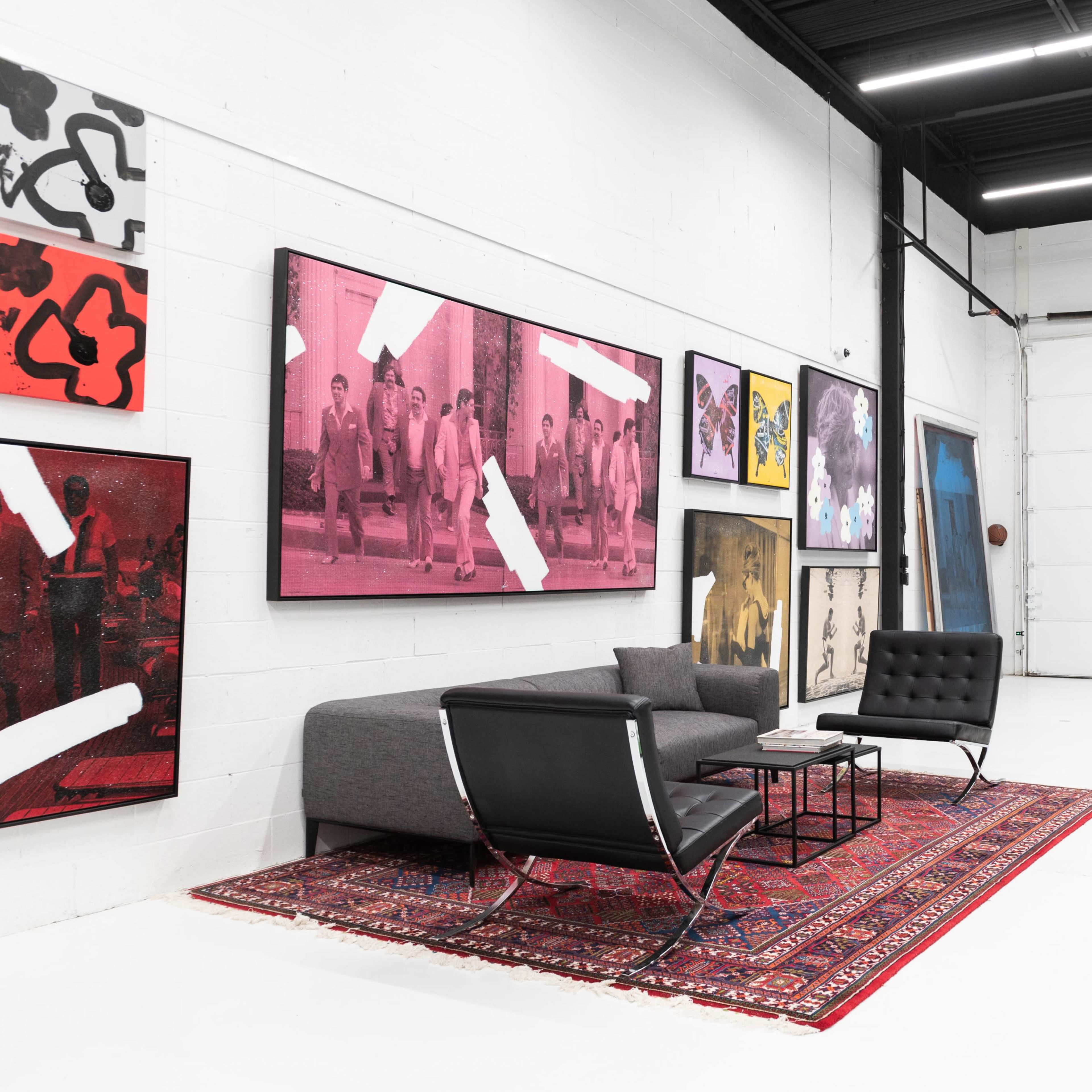 A modern art gallery space with a white floor, featuring various large framed artworks on the walls and a minimalist seating area with a gray sofa and a coffee table on a patterned rug.