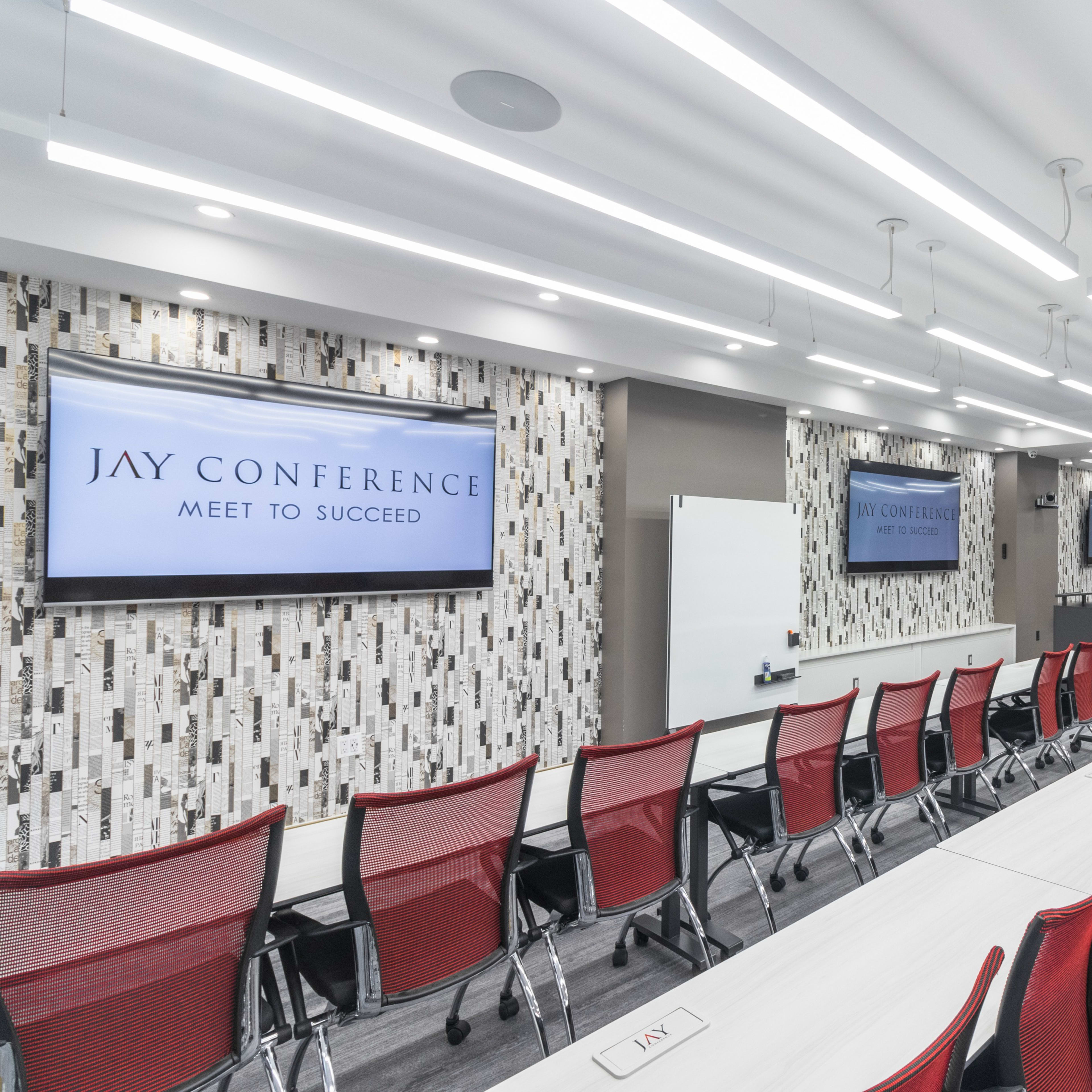 A modern conference room with red chairs arranged around long tables and two large screens displaying "JAY CONFERENCE."
