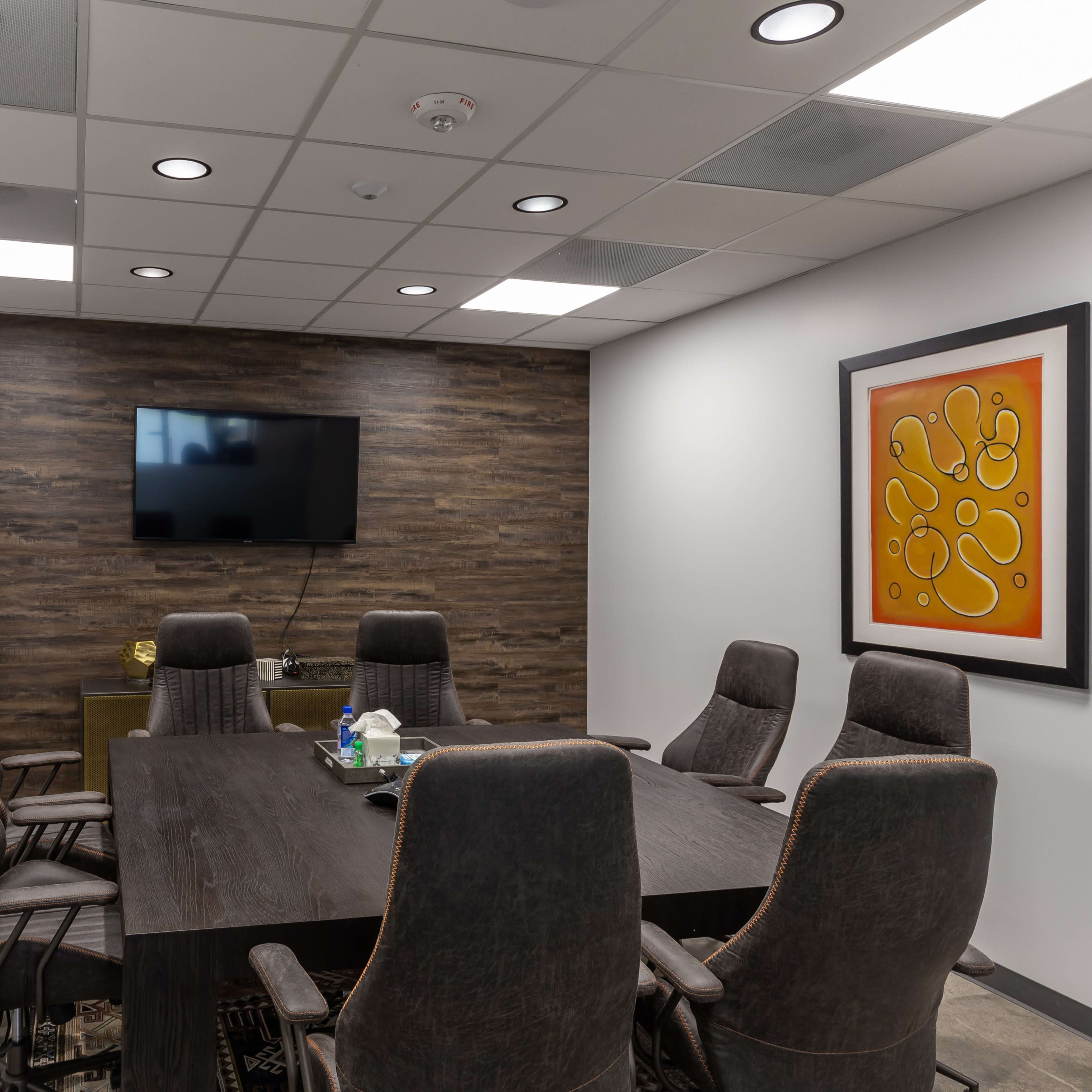 A modern conference room featuring a large wooden table surrounded by eight upholstered chairs, a wall-mounted television, and a whiteboard.