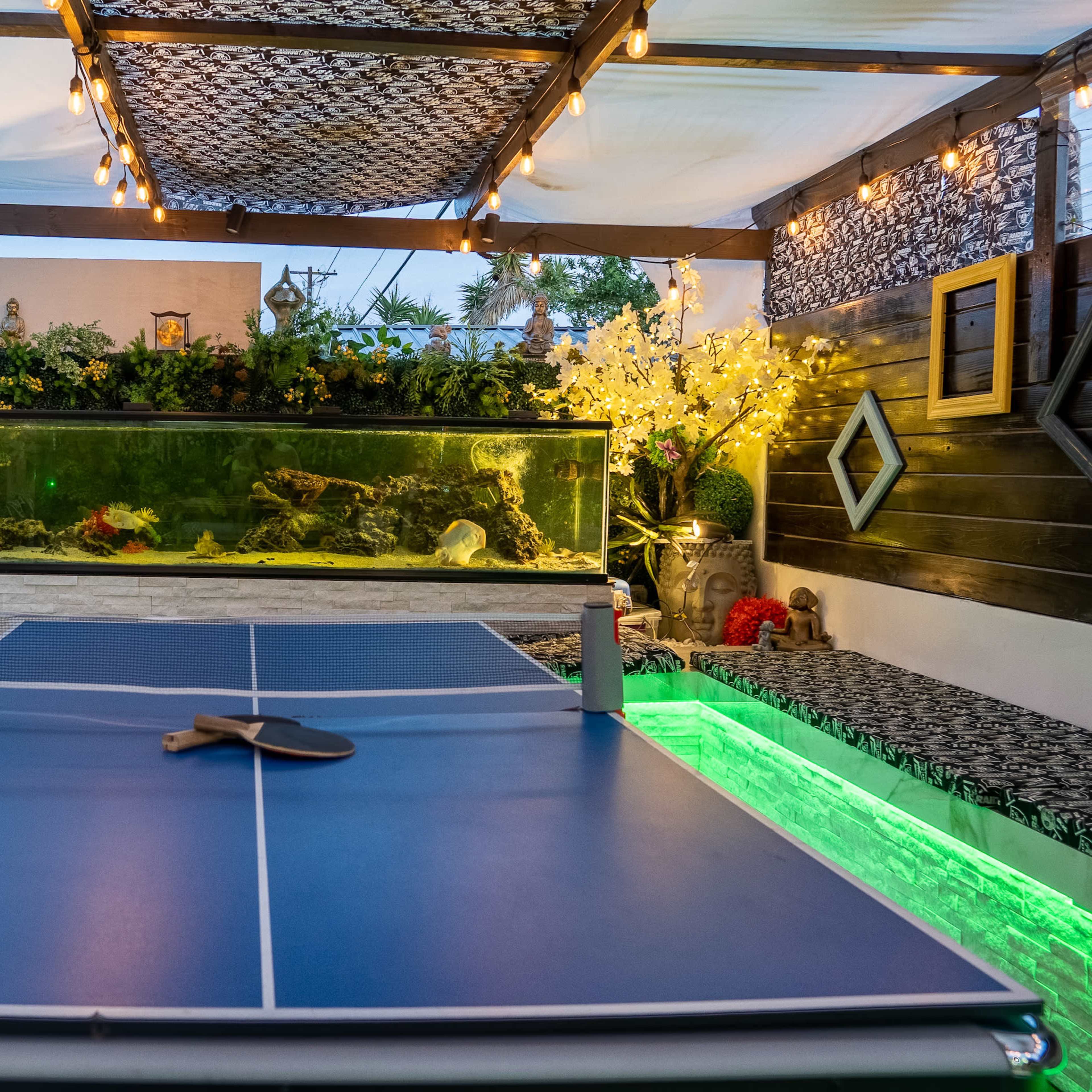 A backyard patio area with a blue ping pong table in the foreground, an aquarium filled with aquatic life in the background, and decorative lighting and plants around the space.