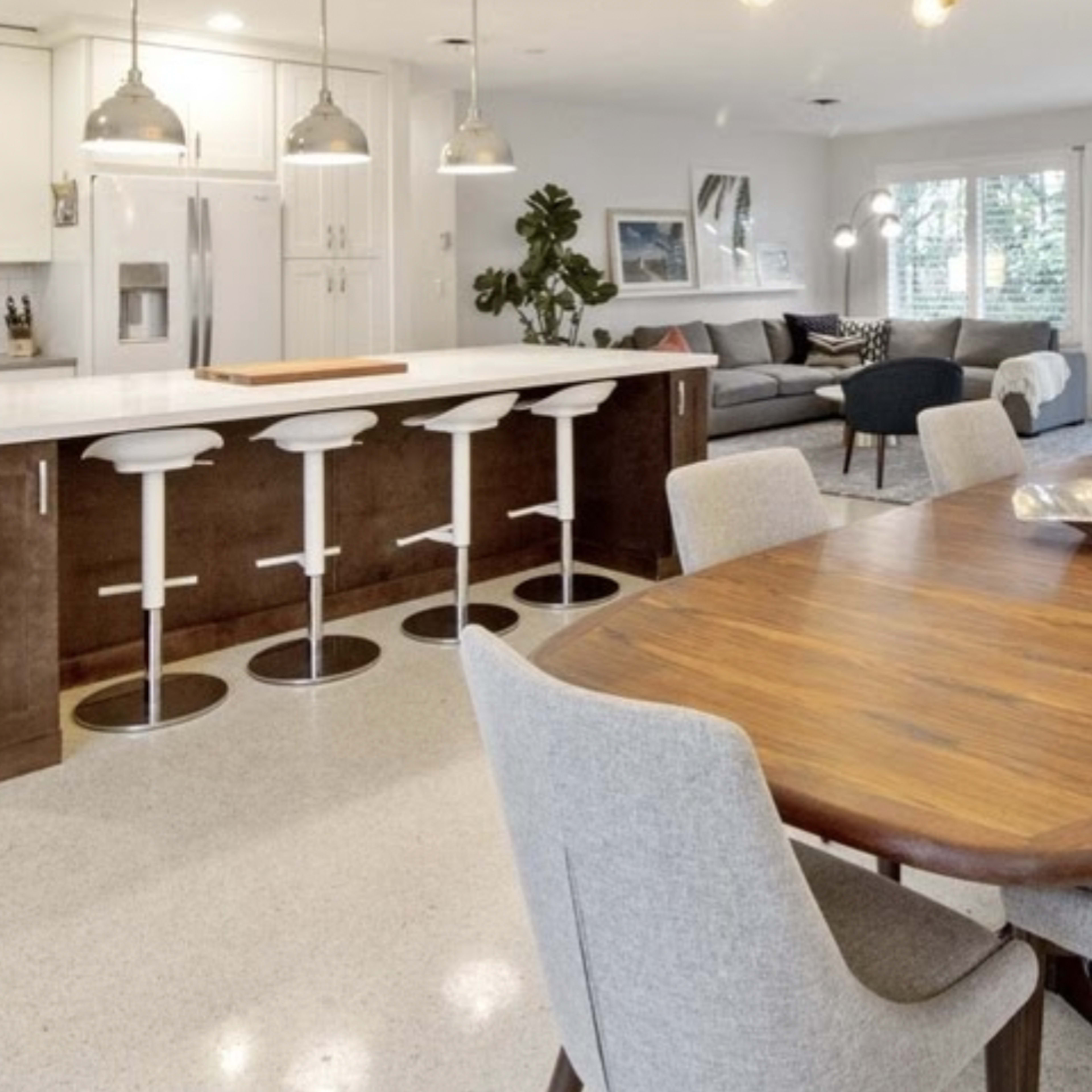 A modern kitchen and dining area featuring a large wooden table surrounded by gray chairs, an island with bar stools, and a living room with a couch and large windows.