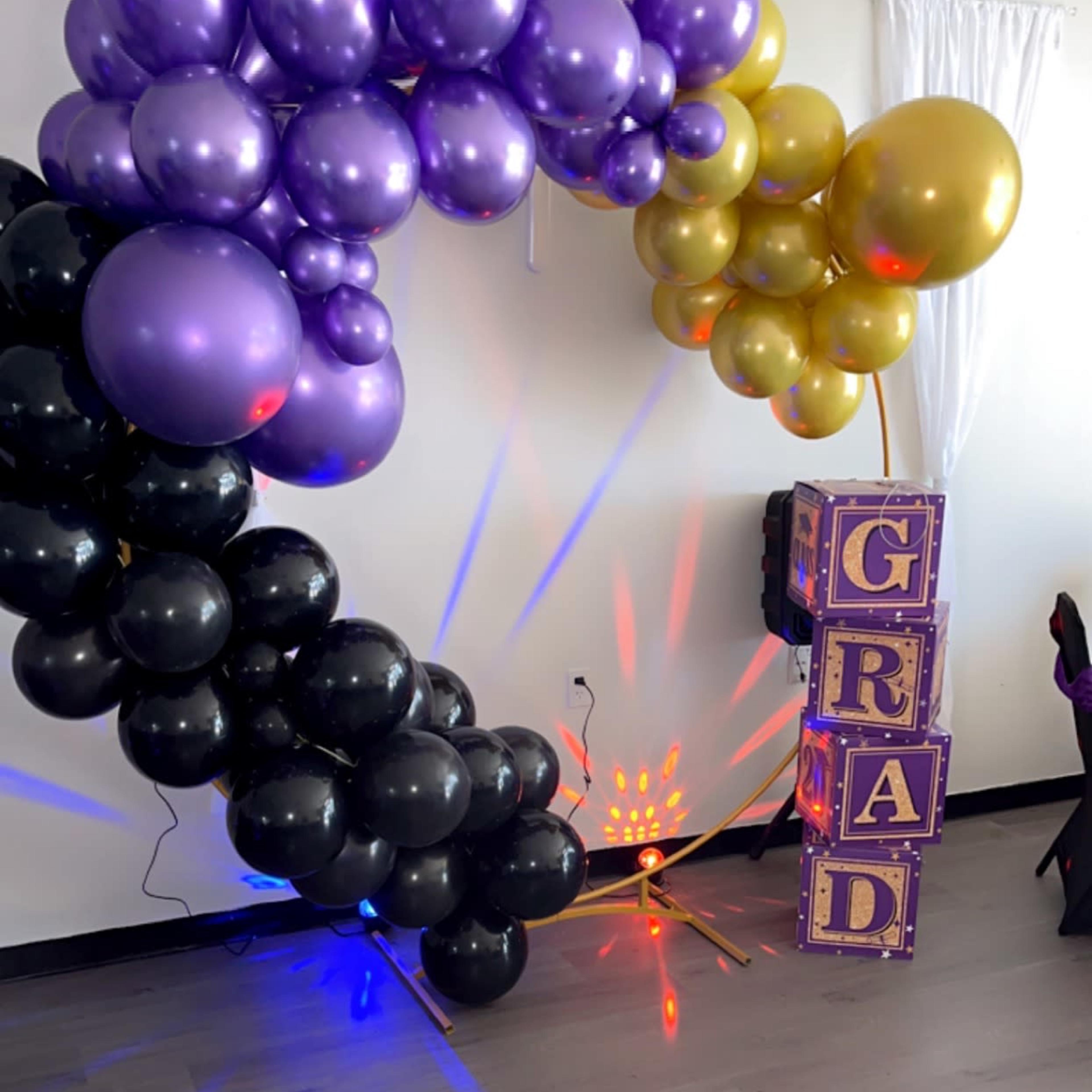 A colorful balloon arch in purple, gold, and black decorates a corner of a room, next to large blocks spelling "GRAD" with lights shining in the background.