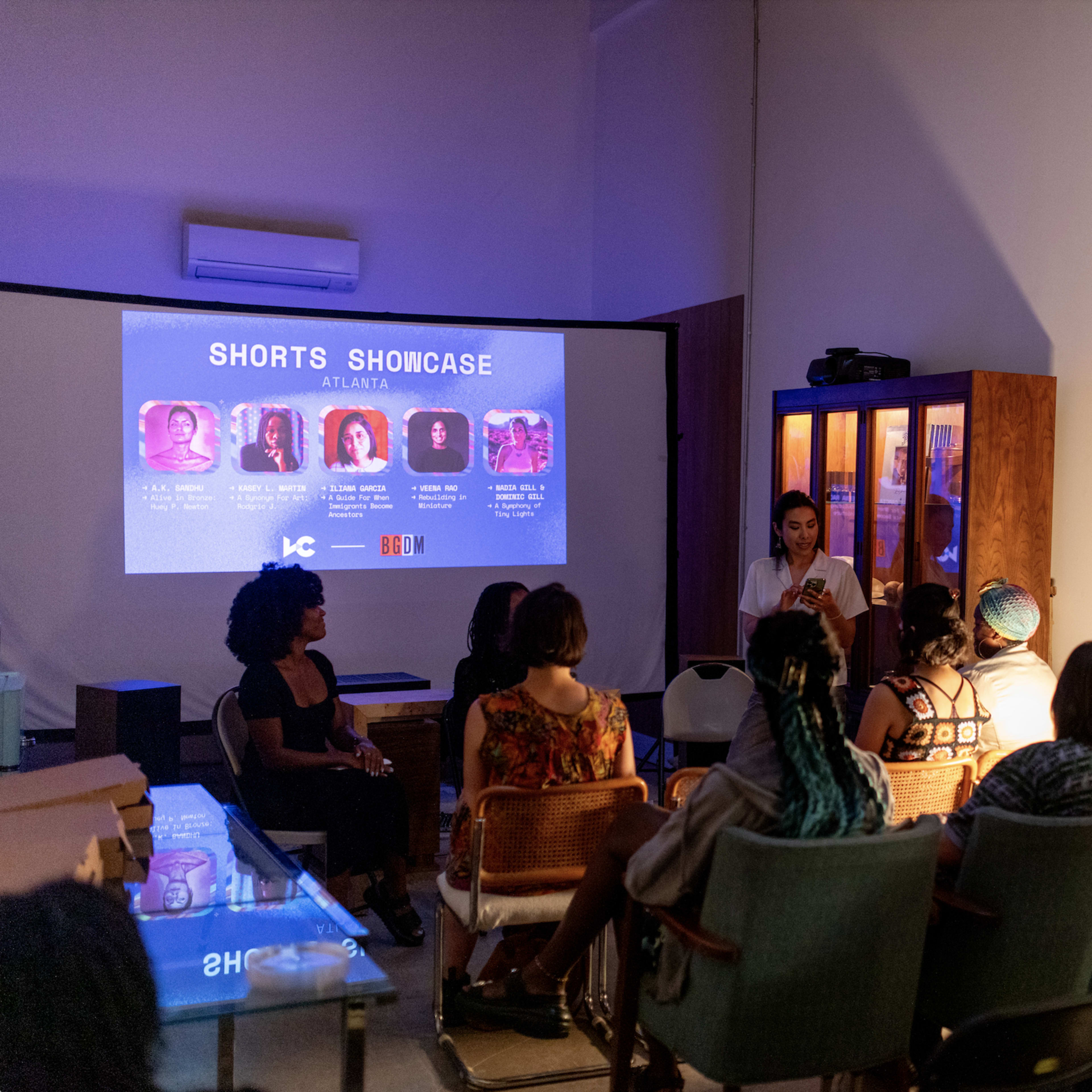 A group of people sits in a room facing a screen displaying information about a shorts showcase event in Atlanta, while a woman stands in front speaking to the audience.
