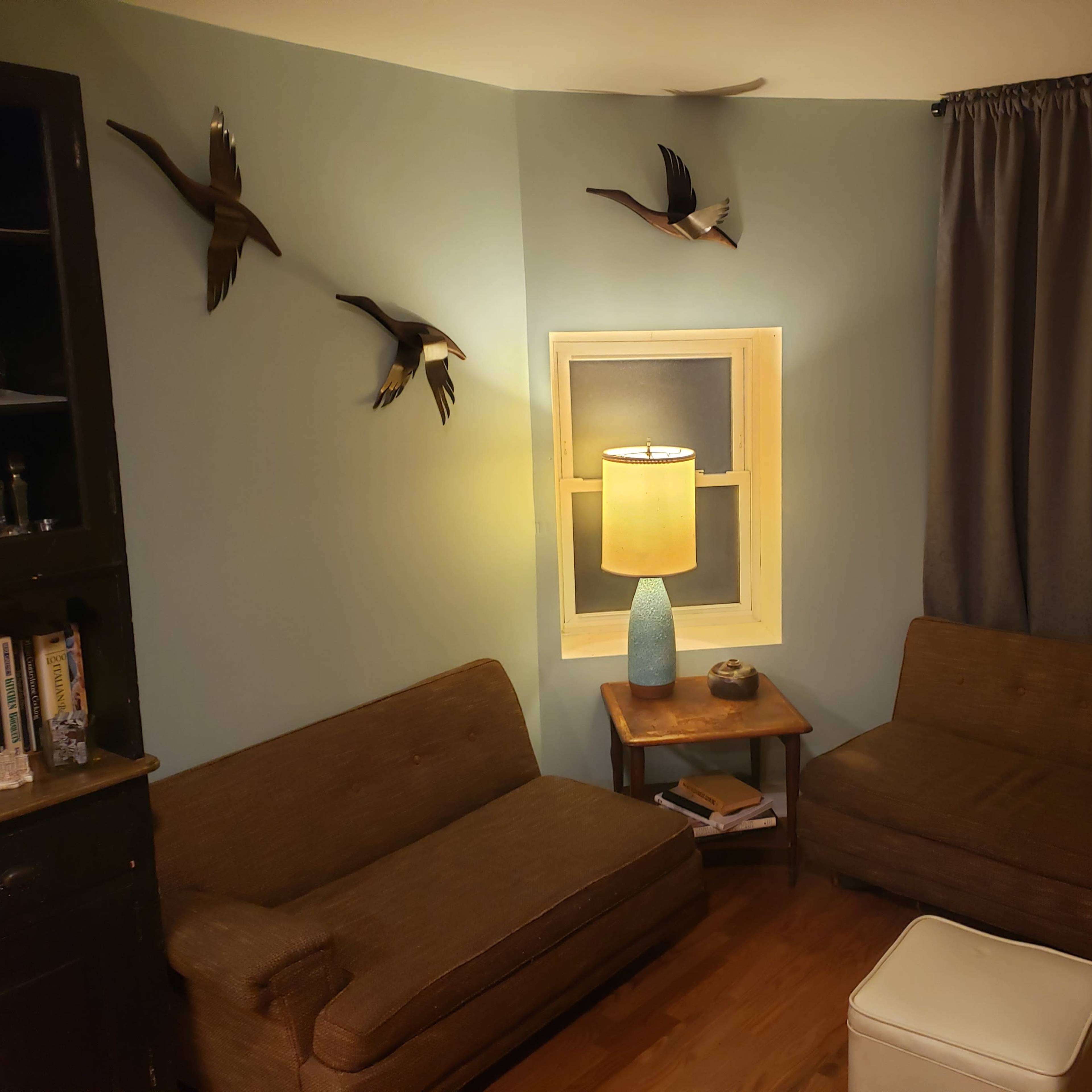 A cozy corner of a living room with two brown couches facing a small wooden table, a lamp, and decorative birds mounted on the wall.