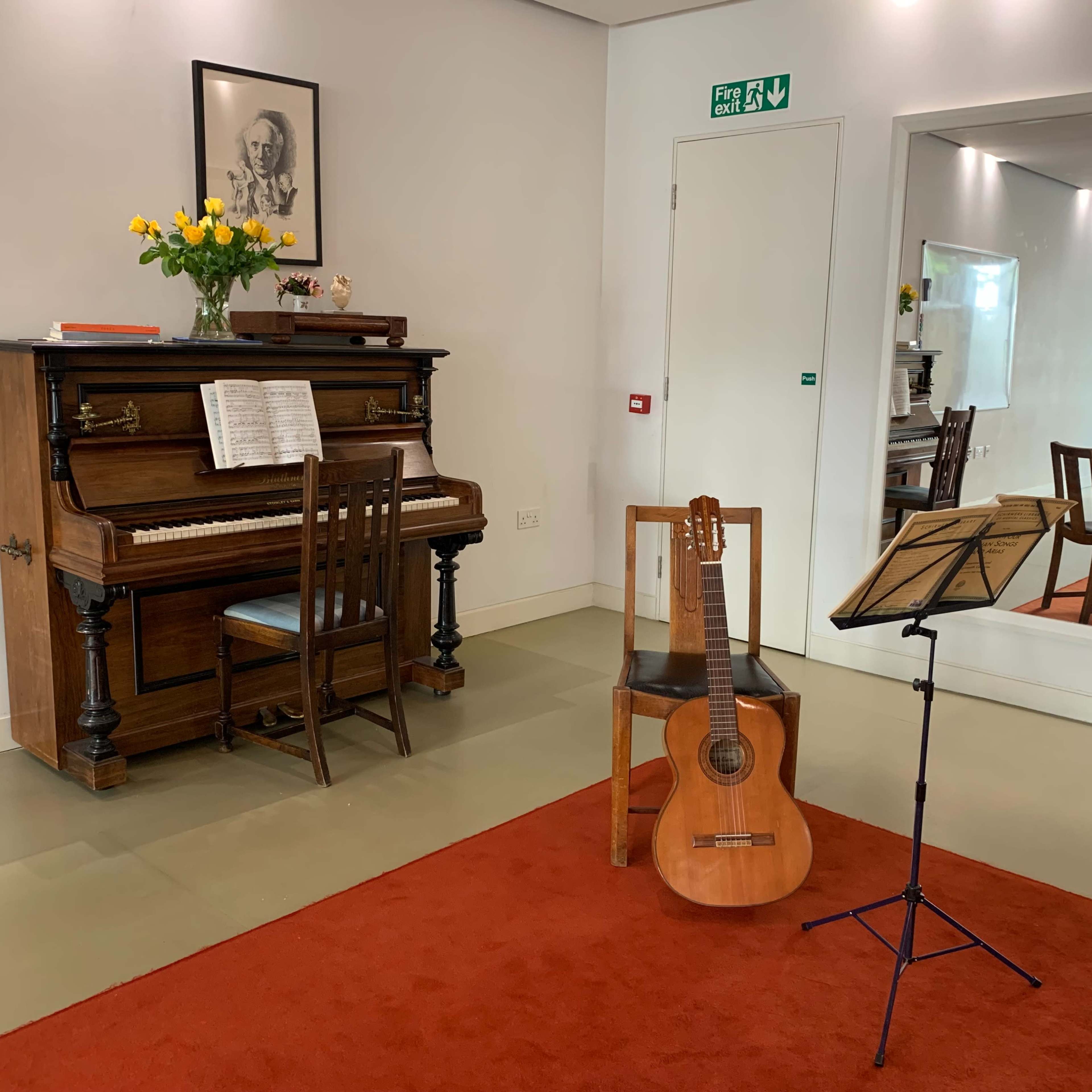 The scene features an upright piano beside a wooden chair, with a guitar and a music stand placed nearby on a red carpet in a well-lit room.