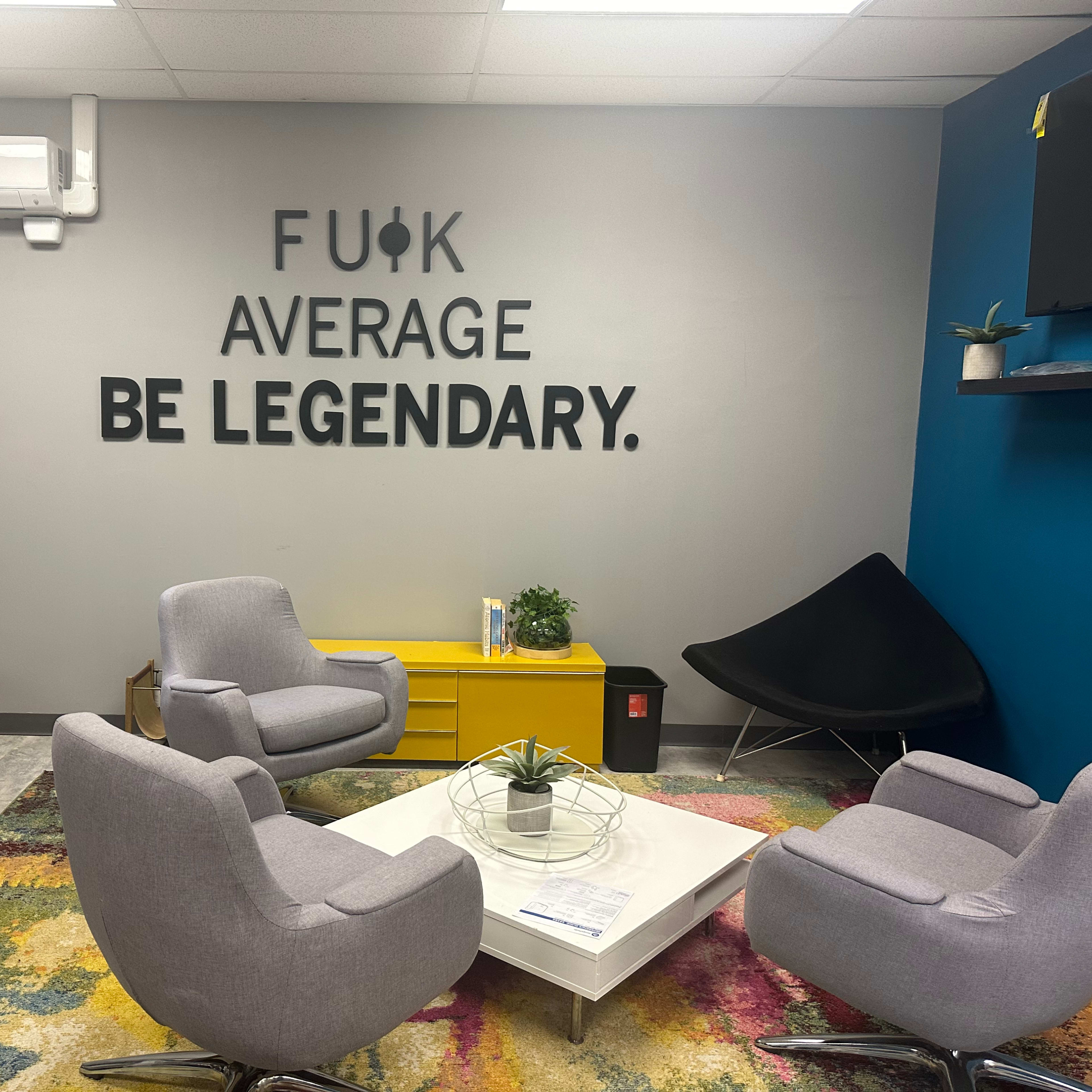A modern office lounge with gray chairs, a round coffee table, a colorful rug, and a bold wall display featuring text that encourages a legendary mindset.