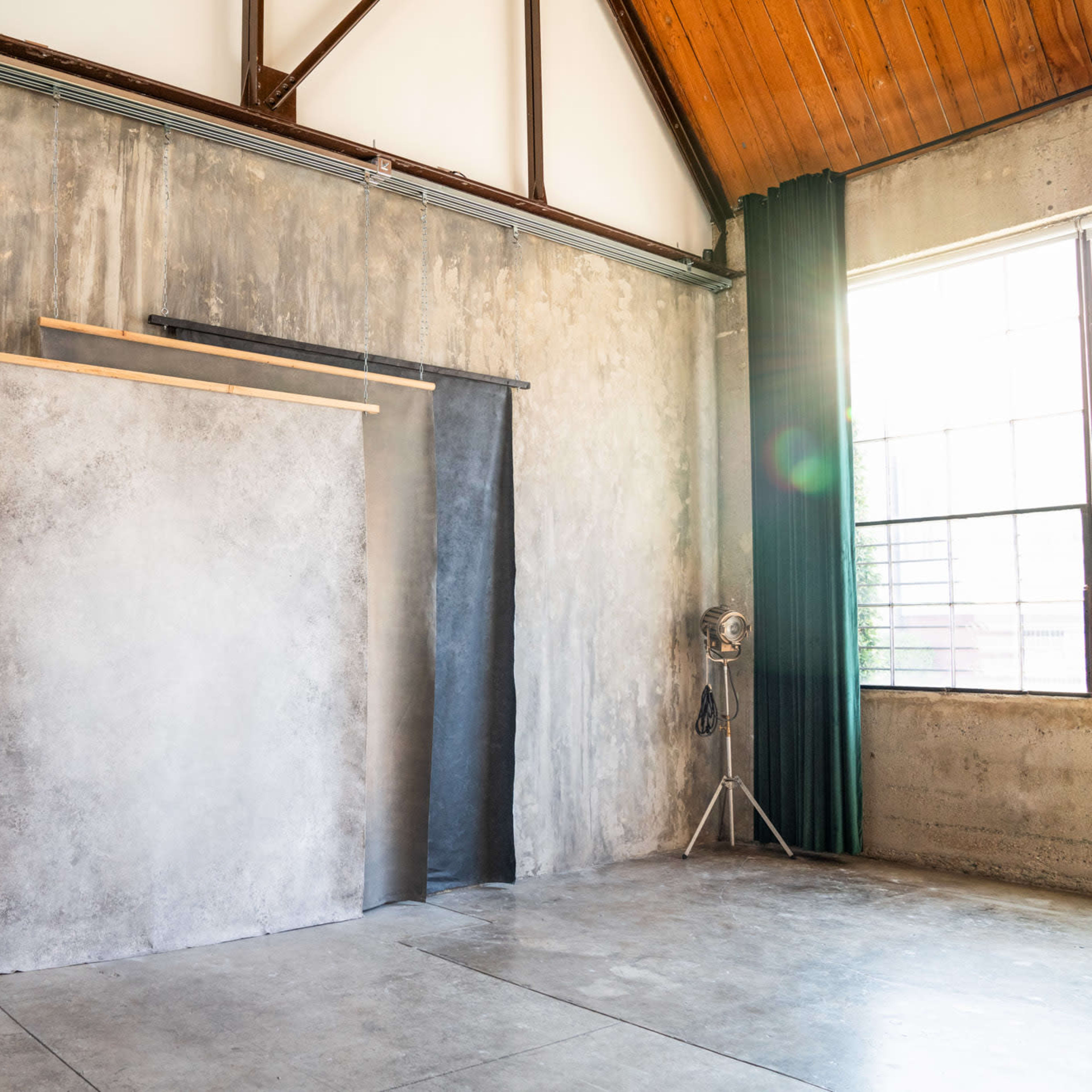 An industrial-style studio with a concrete wall, wooden beams overhead, and three backdrop panels hanging near a large window.