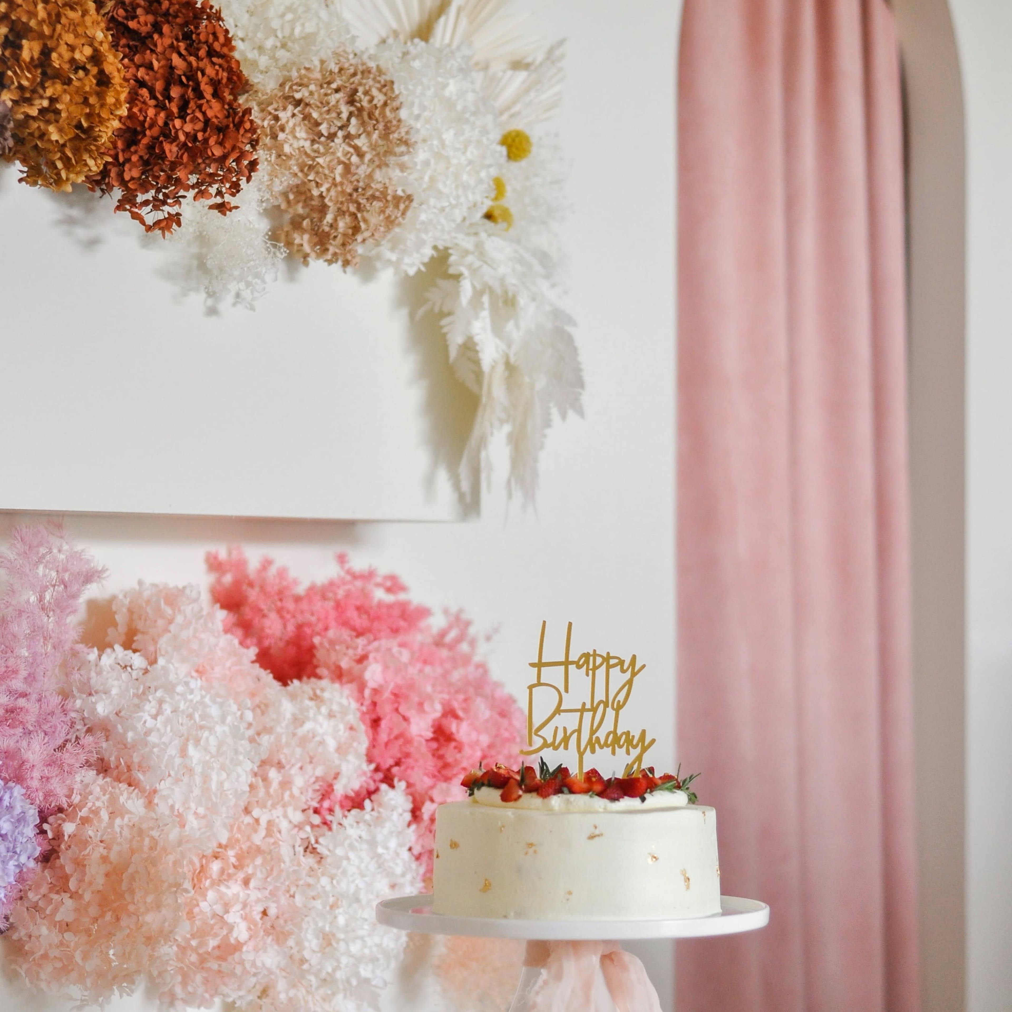 A white birthday cake with a "Happy Birthday" topper is displayed on a pedestal in front of a colorful floral arrangement against a stylish backdrop.