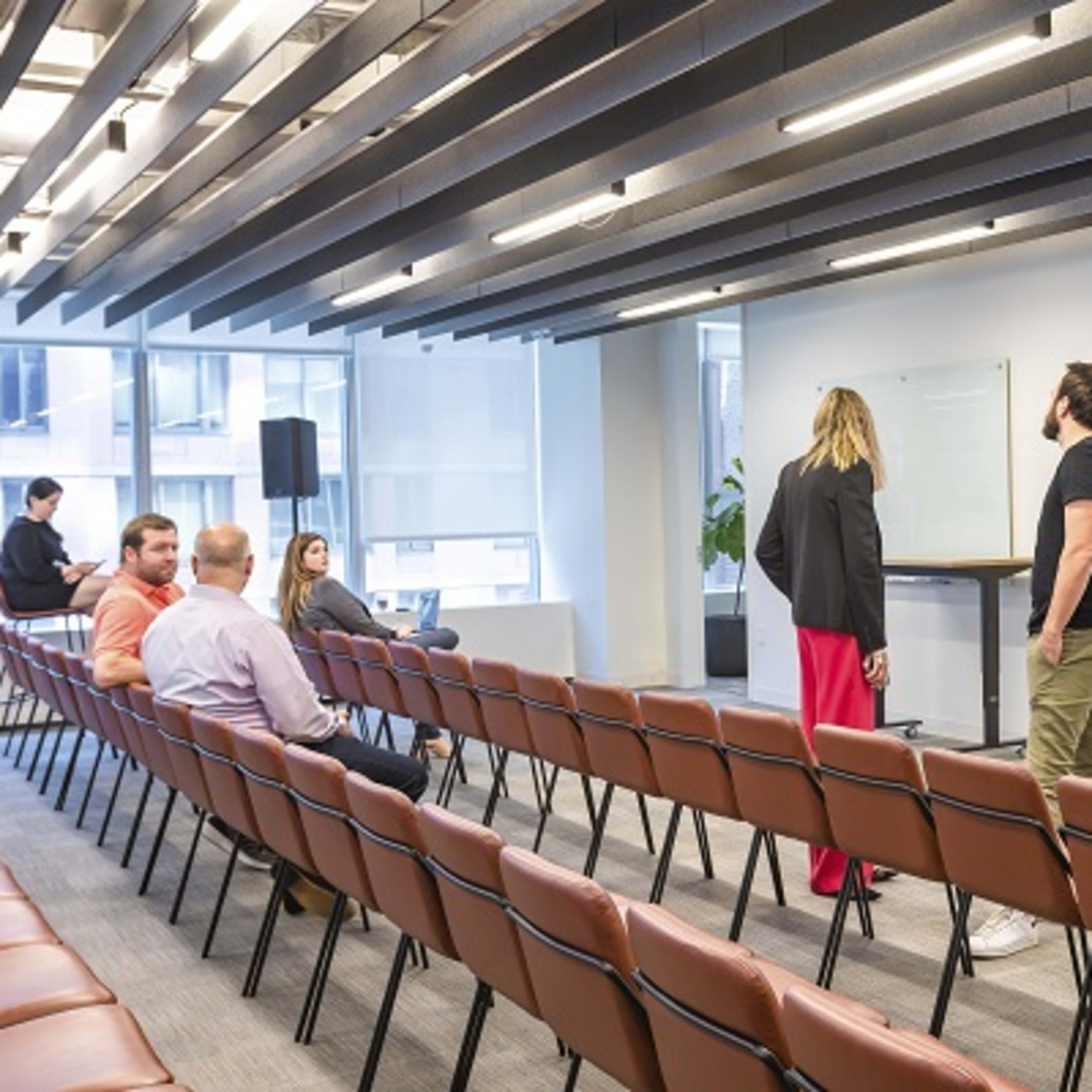 A modern conference room with rows of empty chairs facing a large screen displaying a cityscape, where two people stand near the front and others are seated in the background.