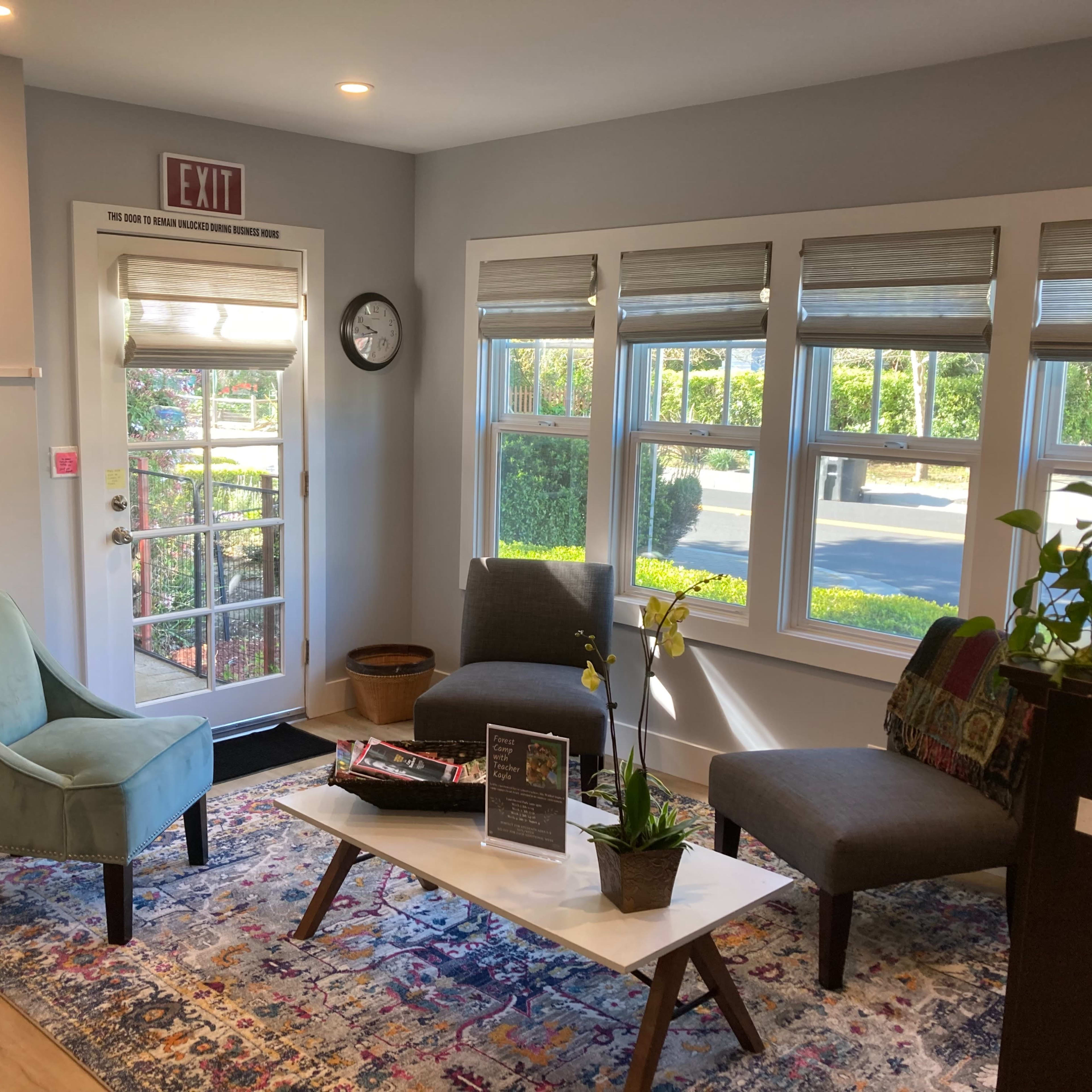 A waiting area with two chairs, a coffee table, a decorative rug, large windows, and a door leading outside.