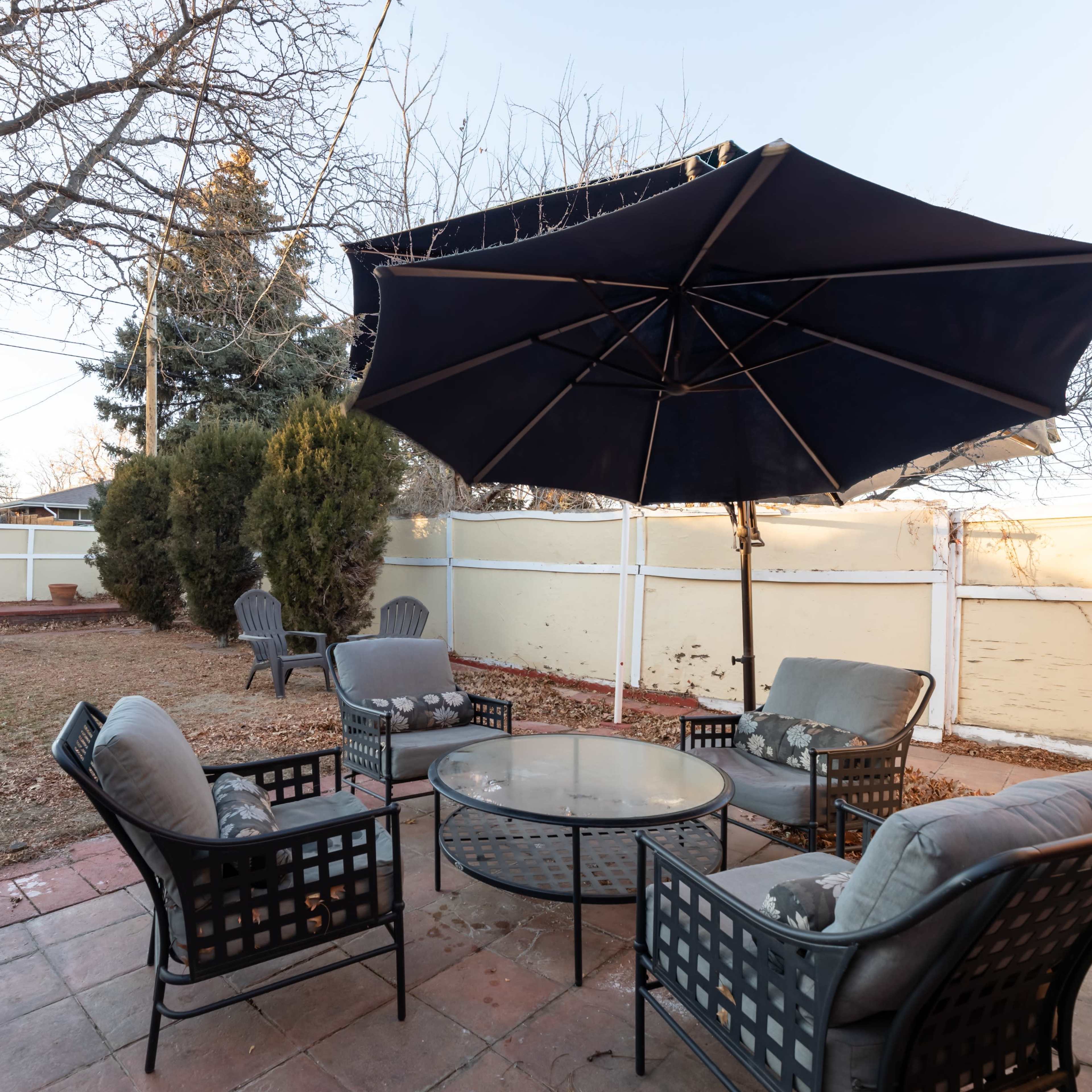 A patio area features a round table surrounded by six chairs, shaded by a large umbrella, with a grassy lawn and trees in the background.