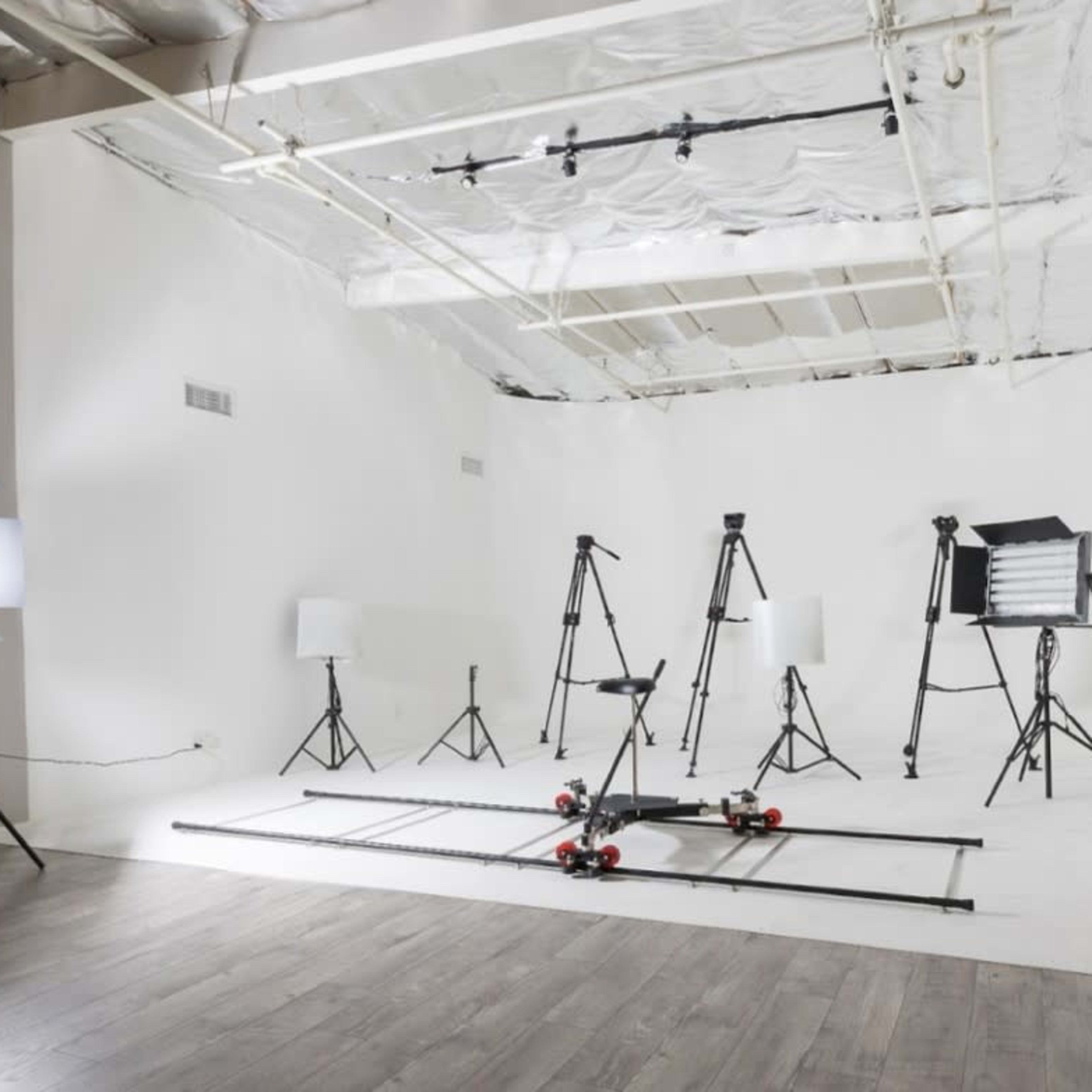 A minimalist studio setup with various photography equipment, including tripods, lighting fixtures, and a track for camera movement, arranged against a white backdrop.