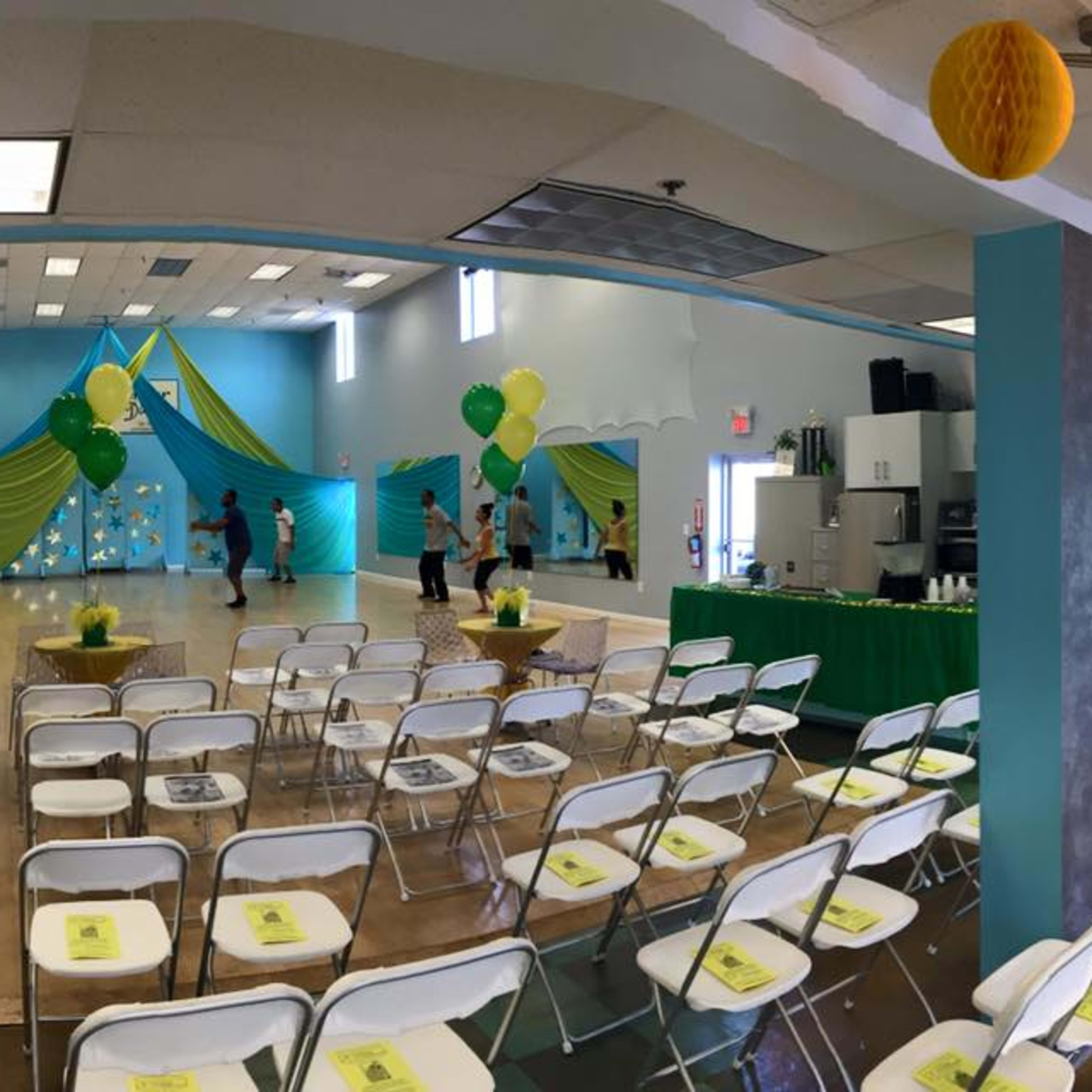 A decorated event space with green and yellow decorations, folding chairs arranged for guests, and a serving area in the background.