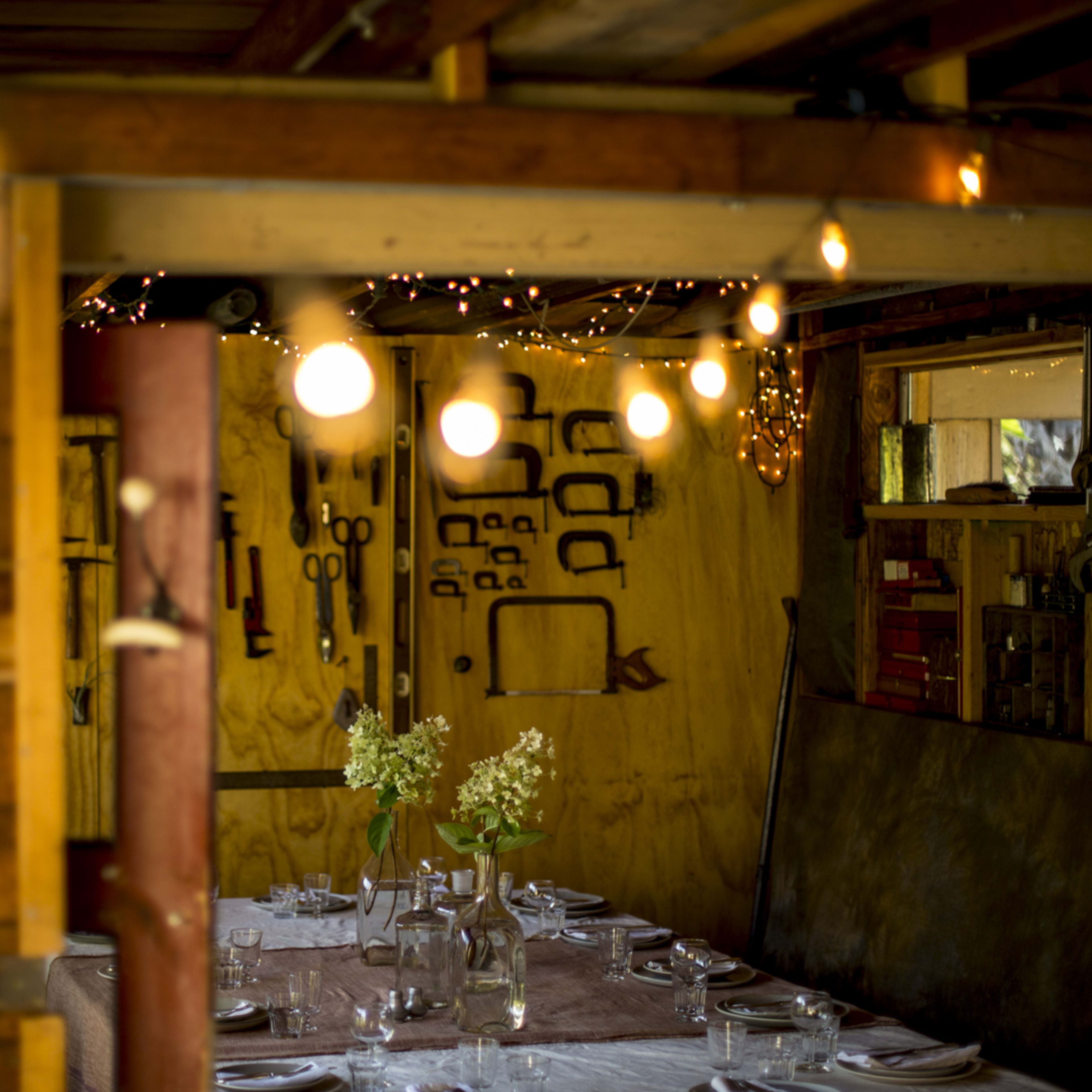 A rustic wooden interior features a dining table set with glassware and flowers, illuminated by string lights overhead.