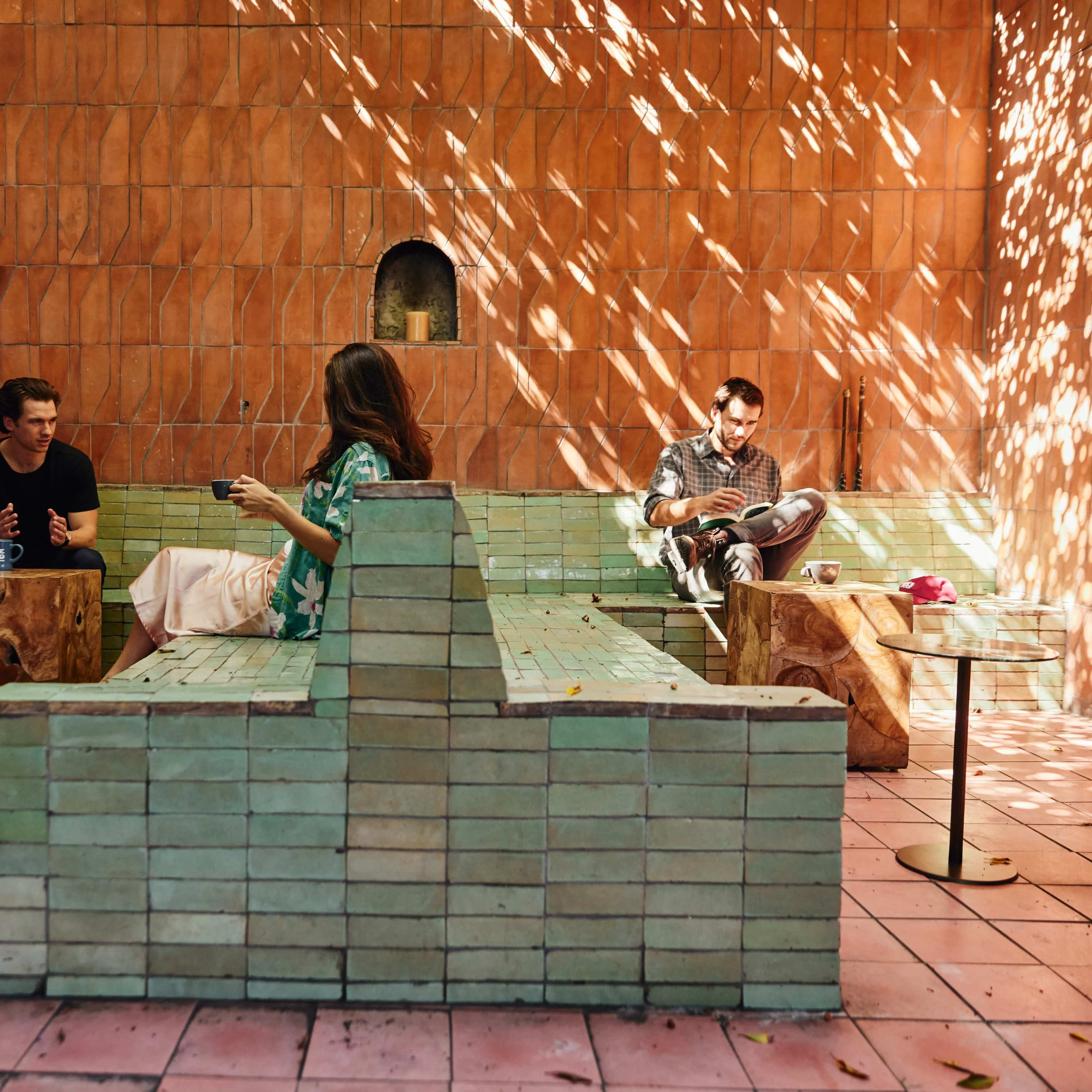 Three people are seated in a courtyard with tiled walls and wooden tables, engaged in conversation and interaction.