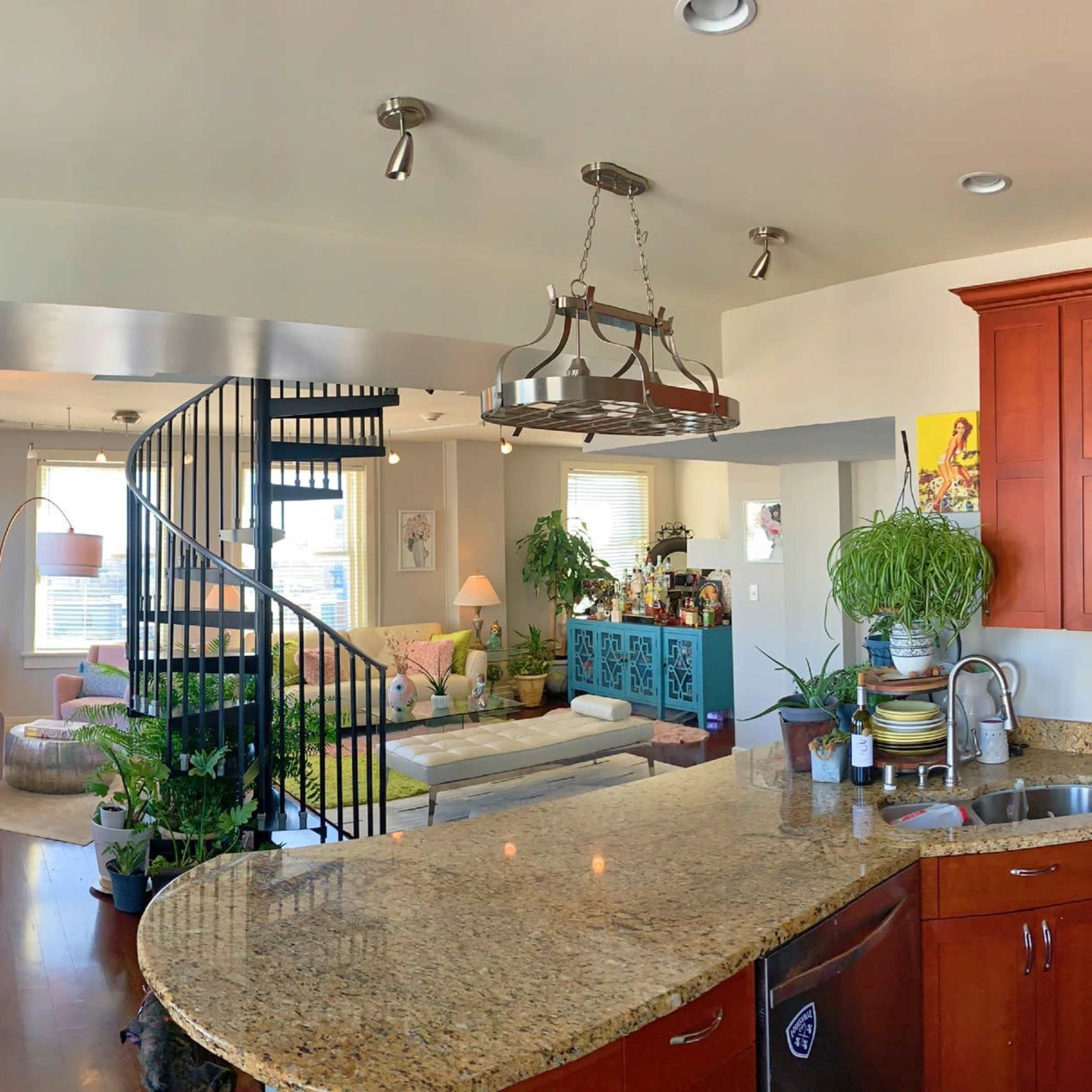 A modern open-concept kitchen with granite countertops and stainless steel appliances, seamlessly connected to a living area featuring a spiral staircase and various plants.