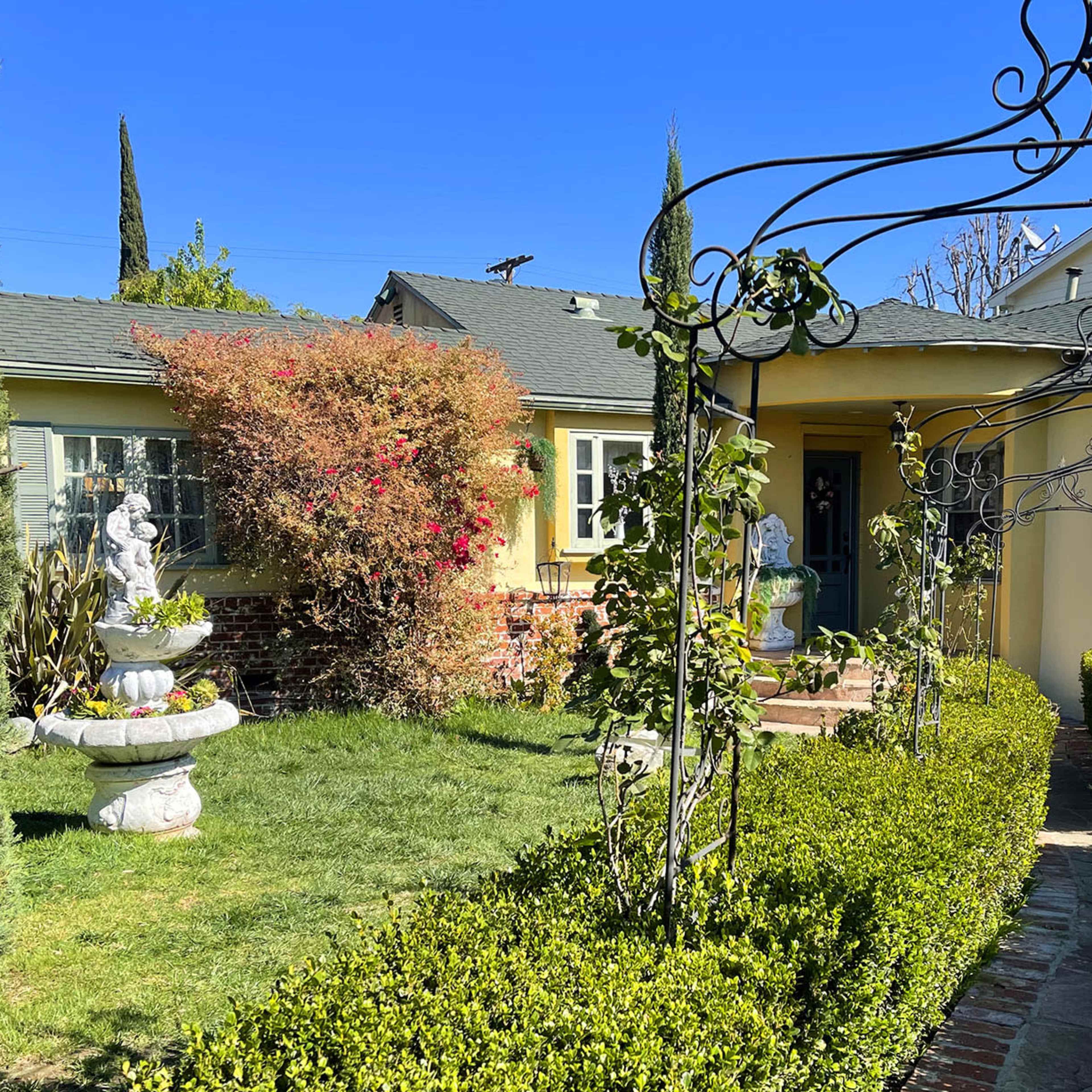 A walkway leads through neatly trimmed hedges to a house with a fountain and decorative statues in the front yard.