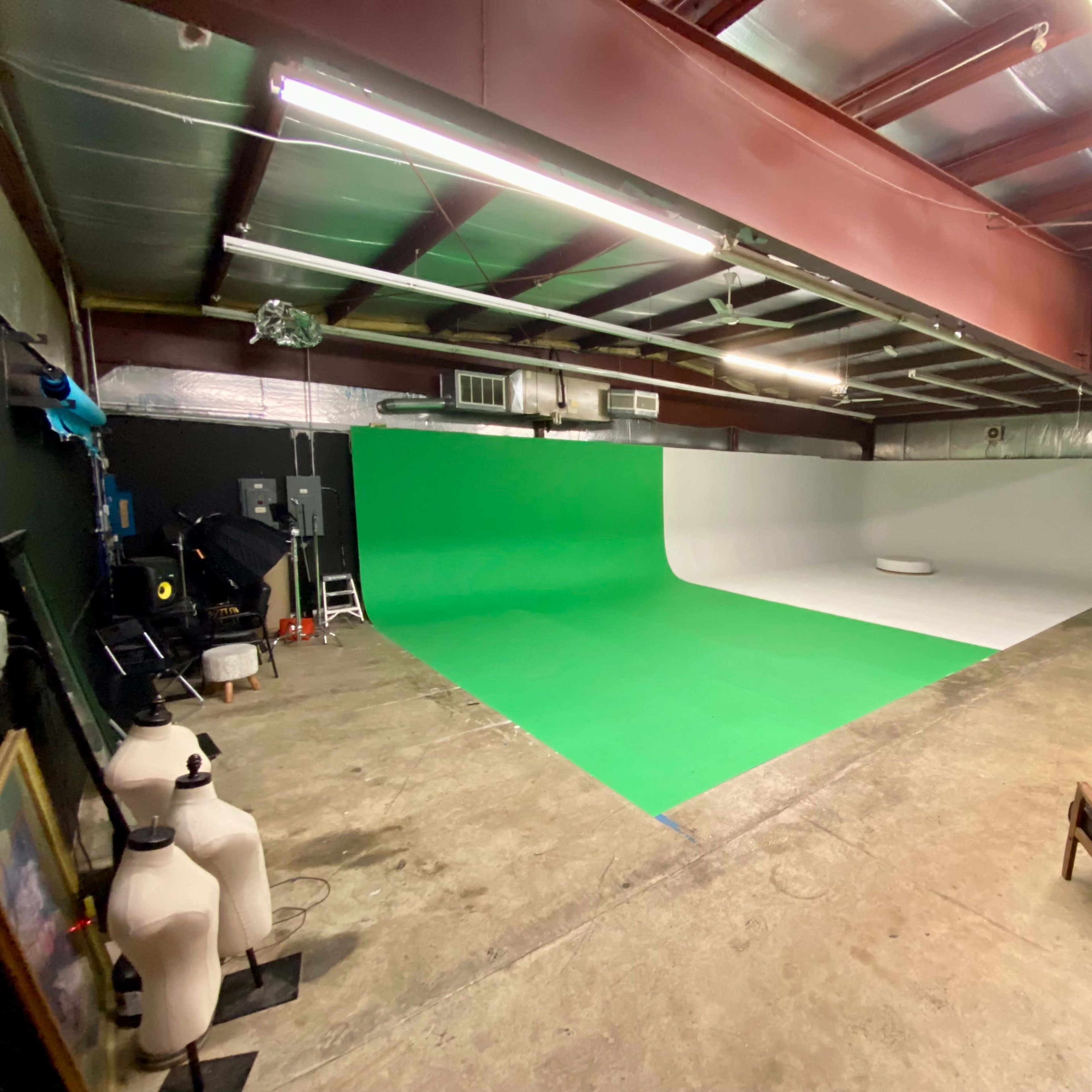 A spacious film studio features a green screen and a white backdrop, with equipment and furniture arranged along the walls.
