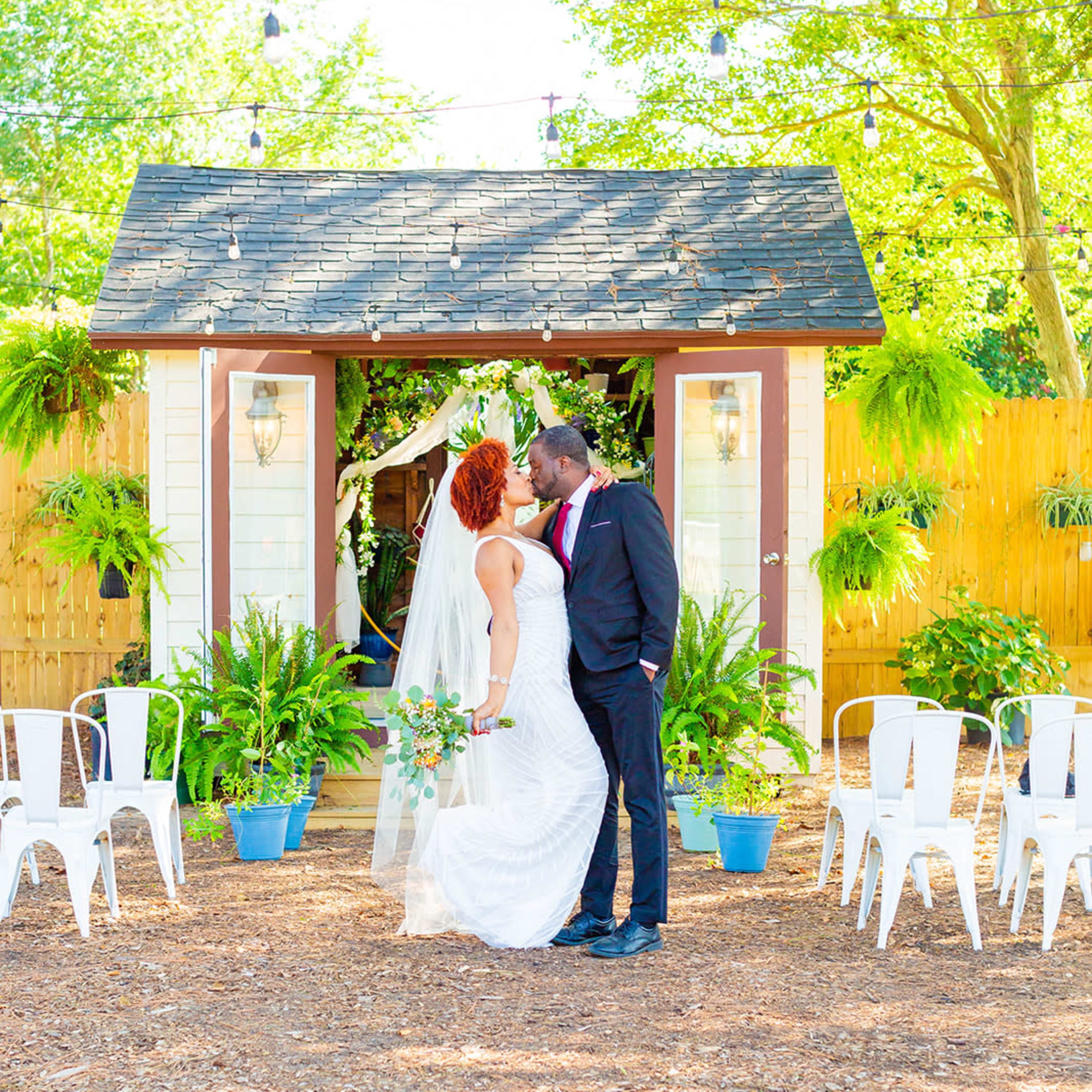 A bride and groom share a kiss under a decorated archway amidst greenery and white chairs in an outdoor wedding setting.