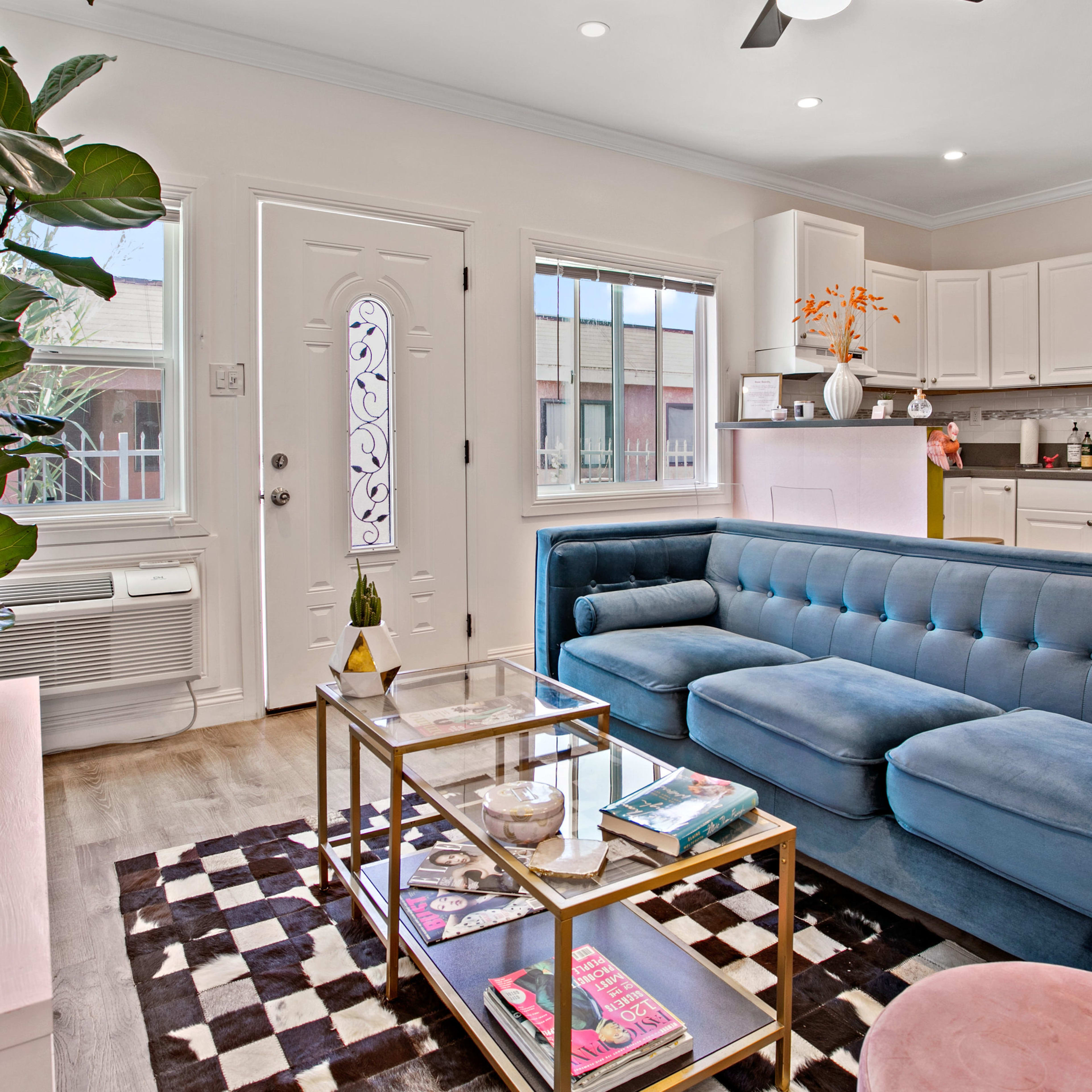 A modern living room features a blue couch, a glass coffee table with magazines, and a white kitchen visible in the background.