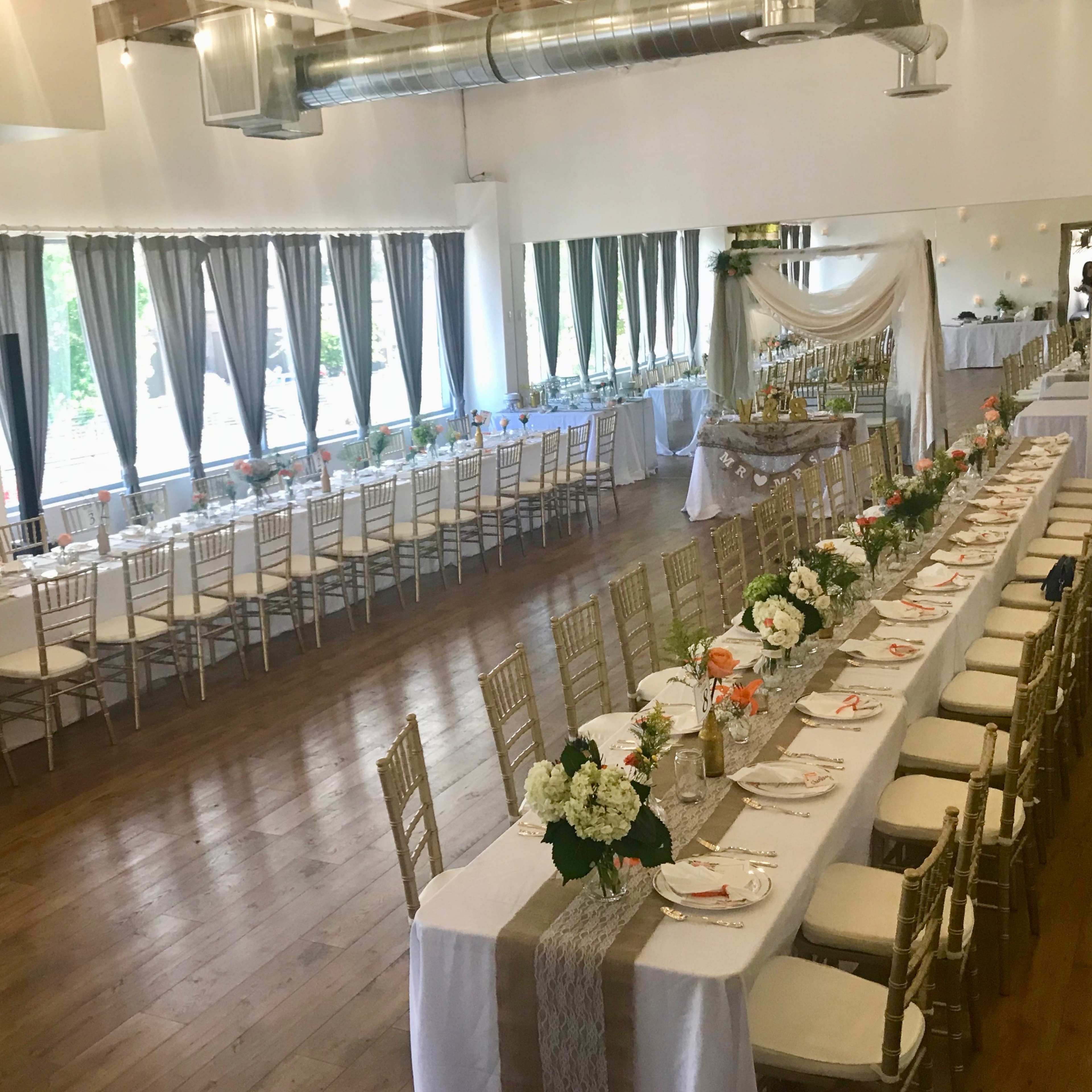 A banquet hall decorated with tables set for a formal event, featuring white tablecloths, floral centerpieces, and large windows with sheer curtains.