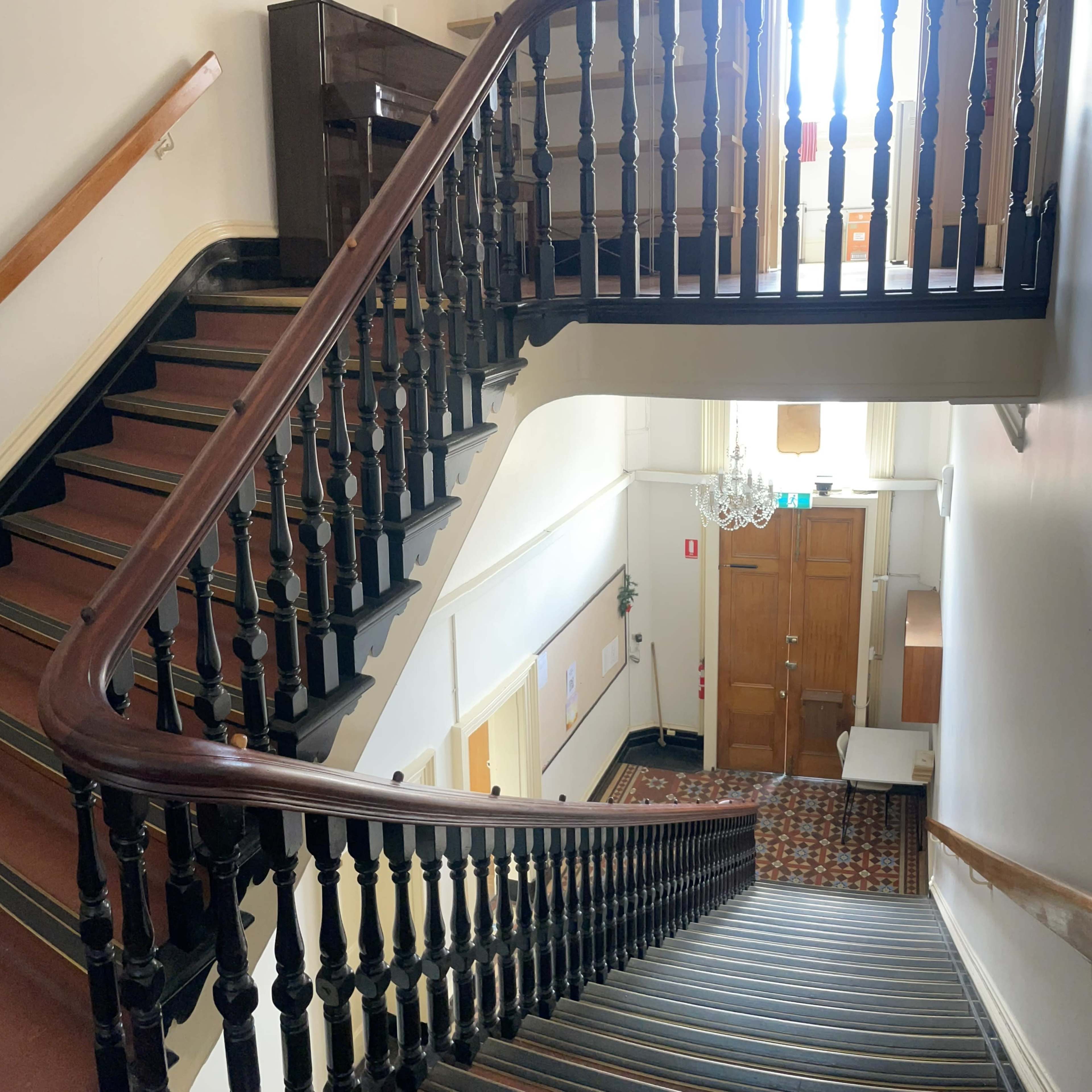 A staircase with a wooden railing leading down to a landing, with a chandelier hanging above and a wooden door at the bottom.