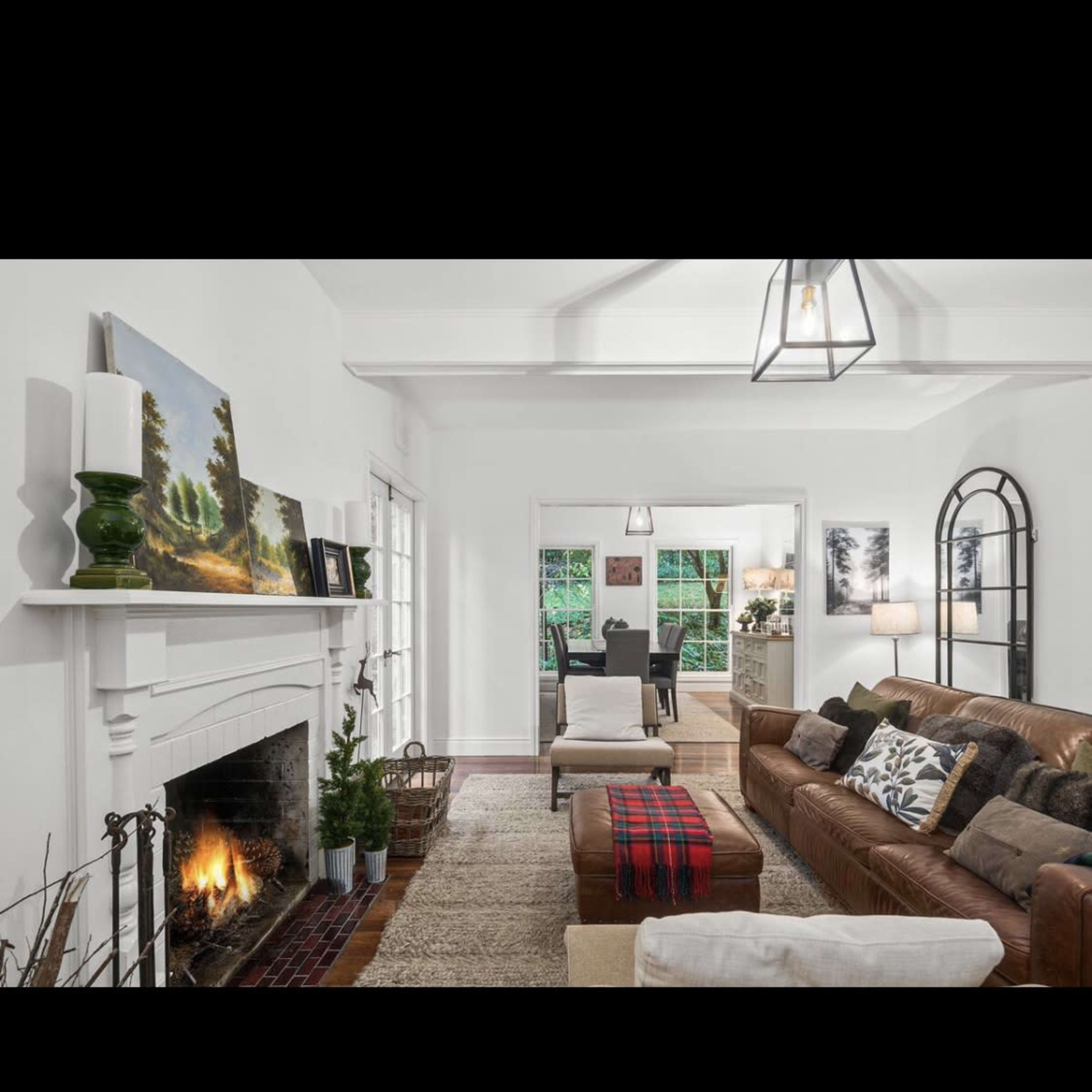 The living room features a brown leather sectional sofa, a coffee table with a red and black plaid throw, a fireplace with a white mantel, and large windows allowing natural light.