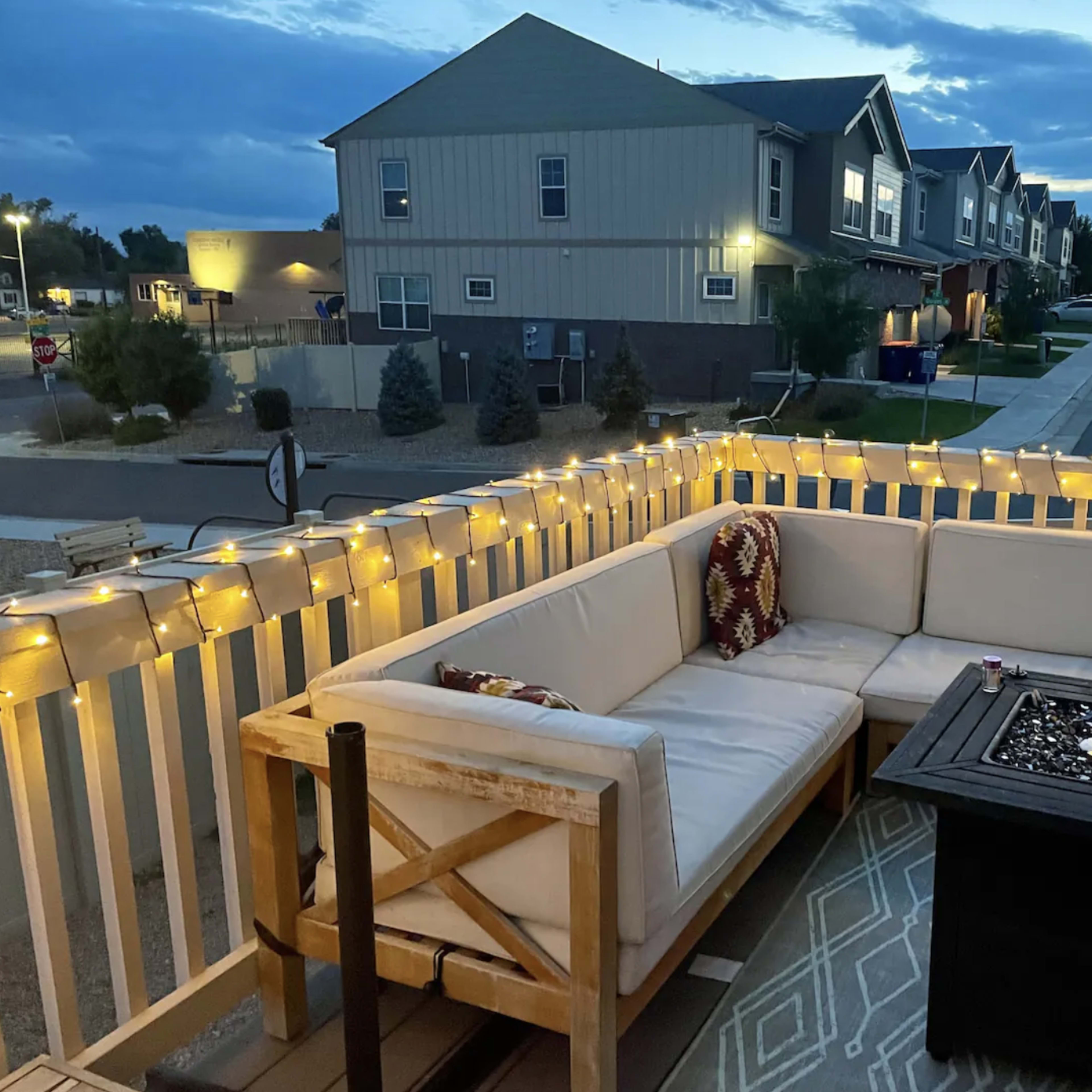 A cozy outdoor seating area is set up on a patio with string lights illuminating a sectional sofa and a fire pit, overlooking a quiet neighborhood at dusk.