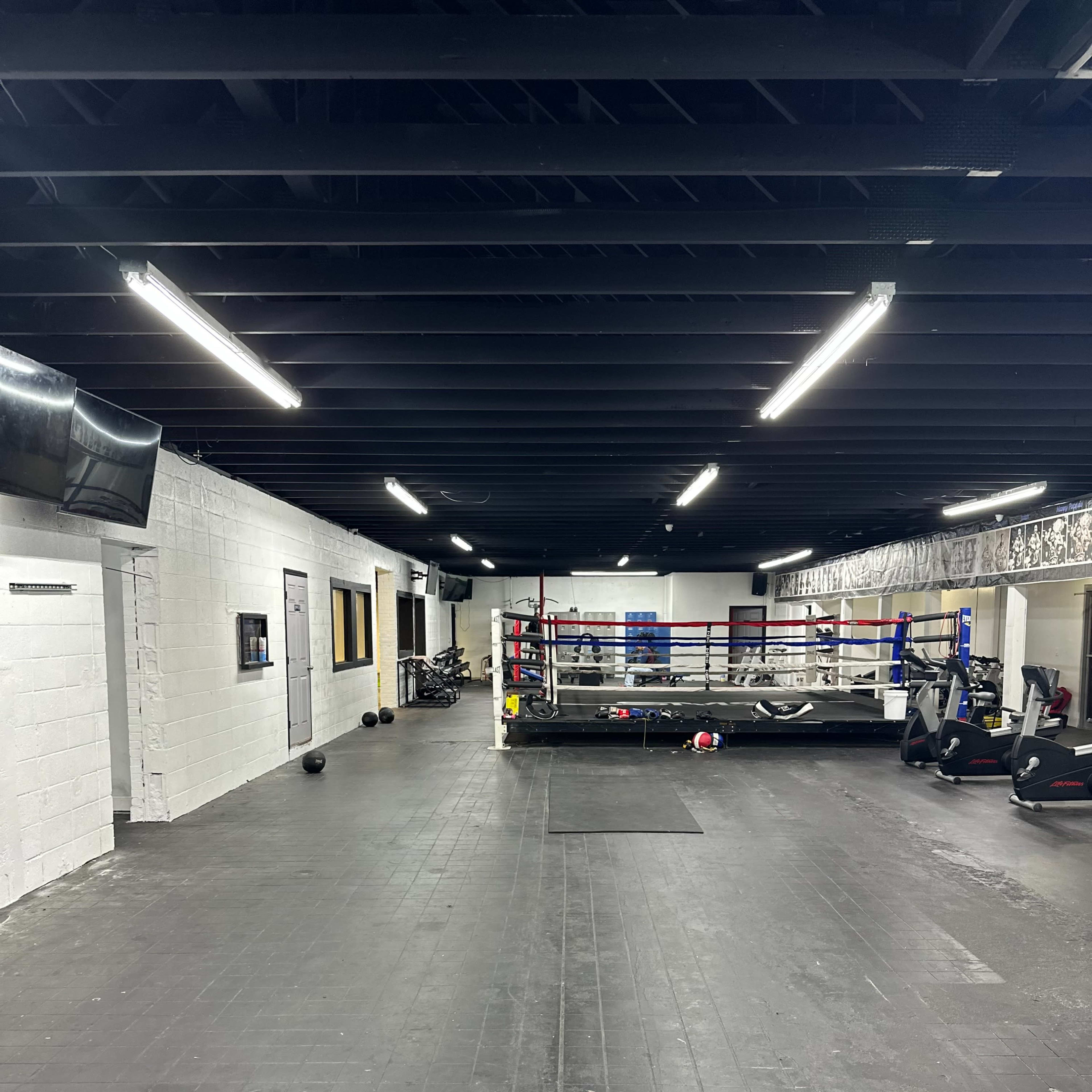 A dimly lit gym with a boxing ring, exercise equipment, and sparsely placed weights along a black rubber floor.