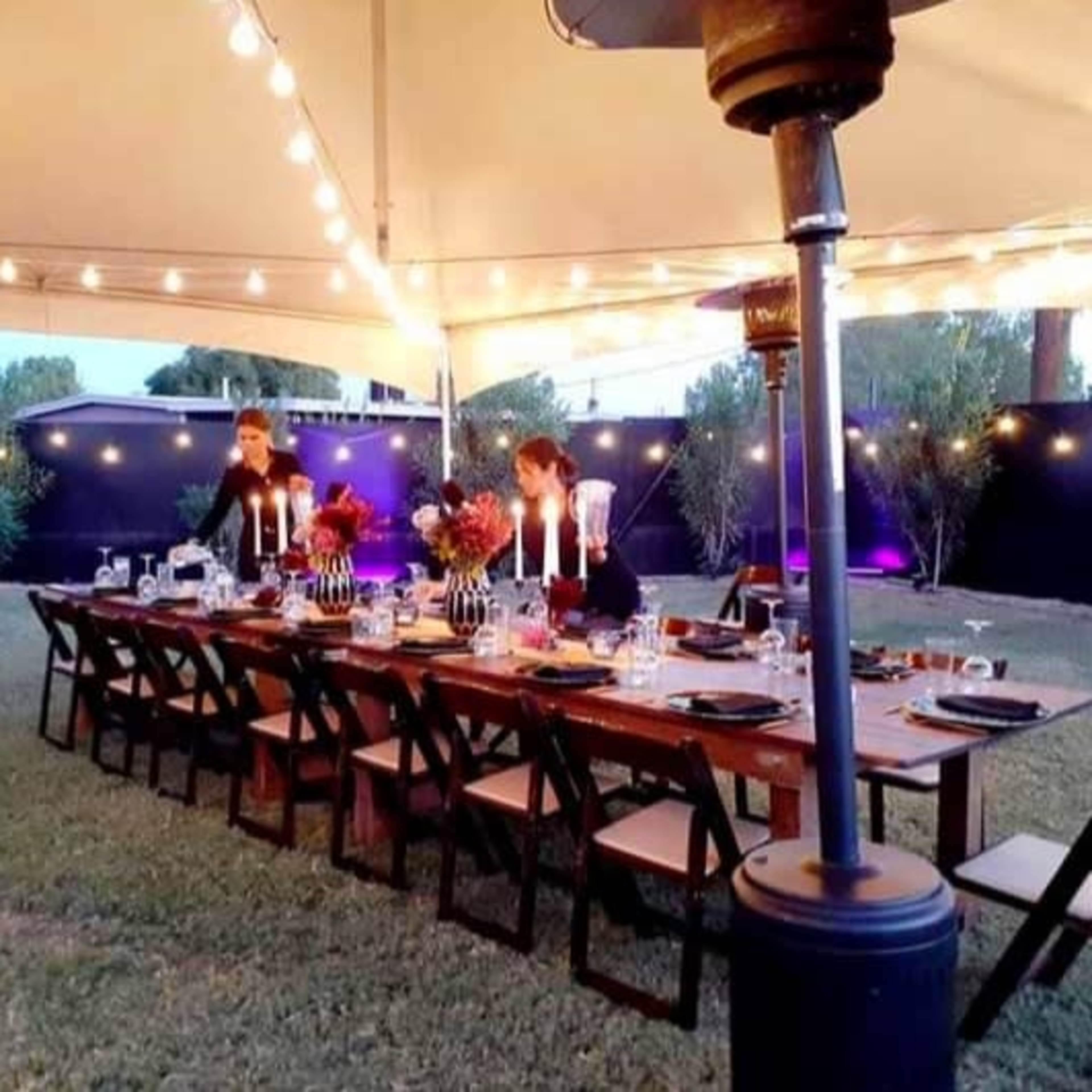 A long wooden dining table is set under a large tent, decorated with candles and flowers, surrounded by chairs and illuminated by string lights.