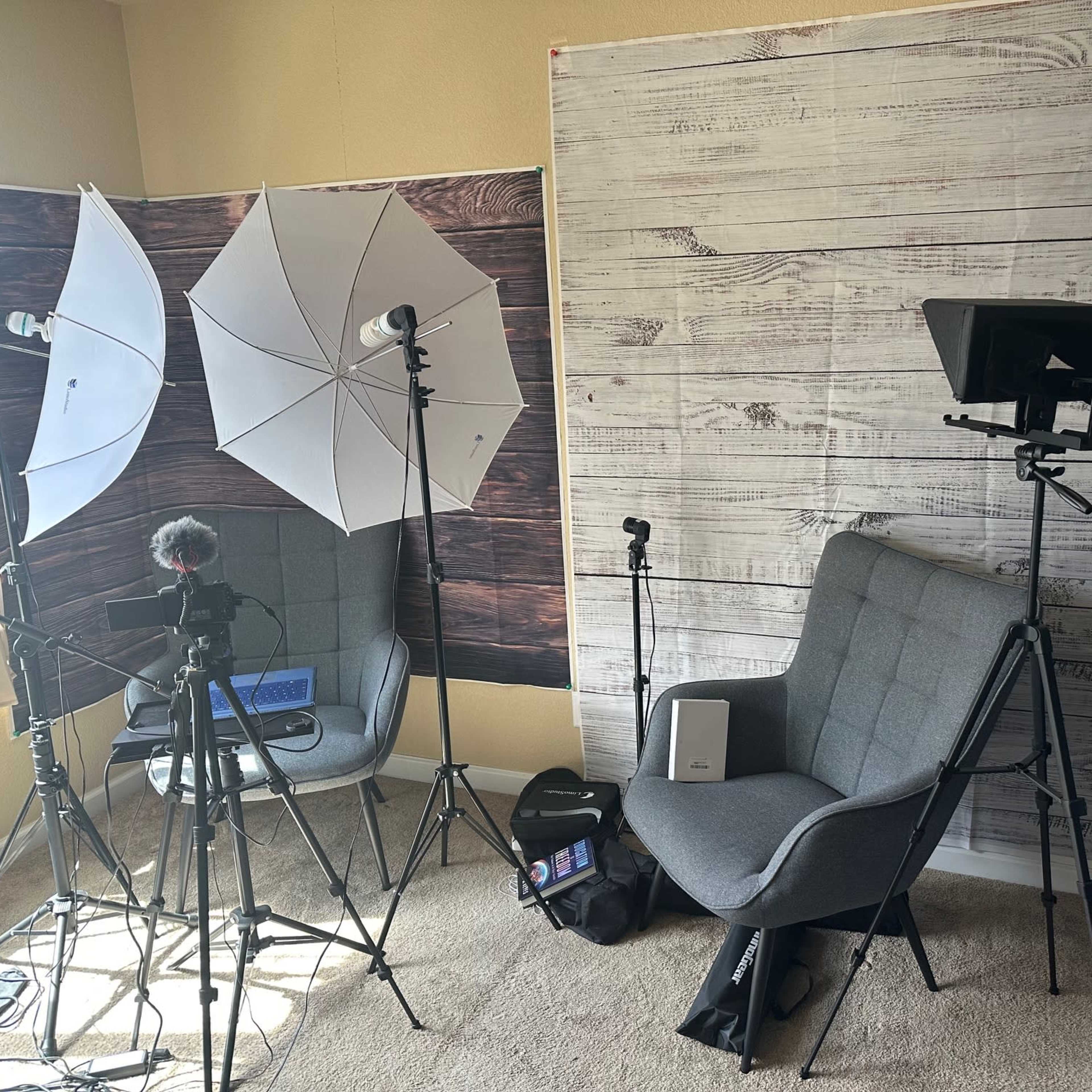A studio setup featuring softbox lights, a camera on a tripod, a gray chair, and a backdrop resembling wooden planks.