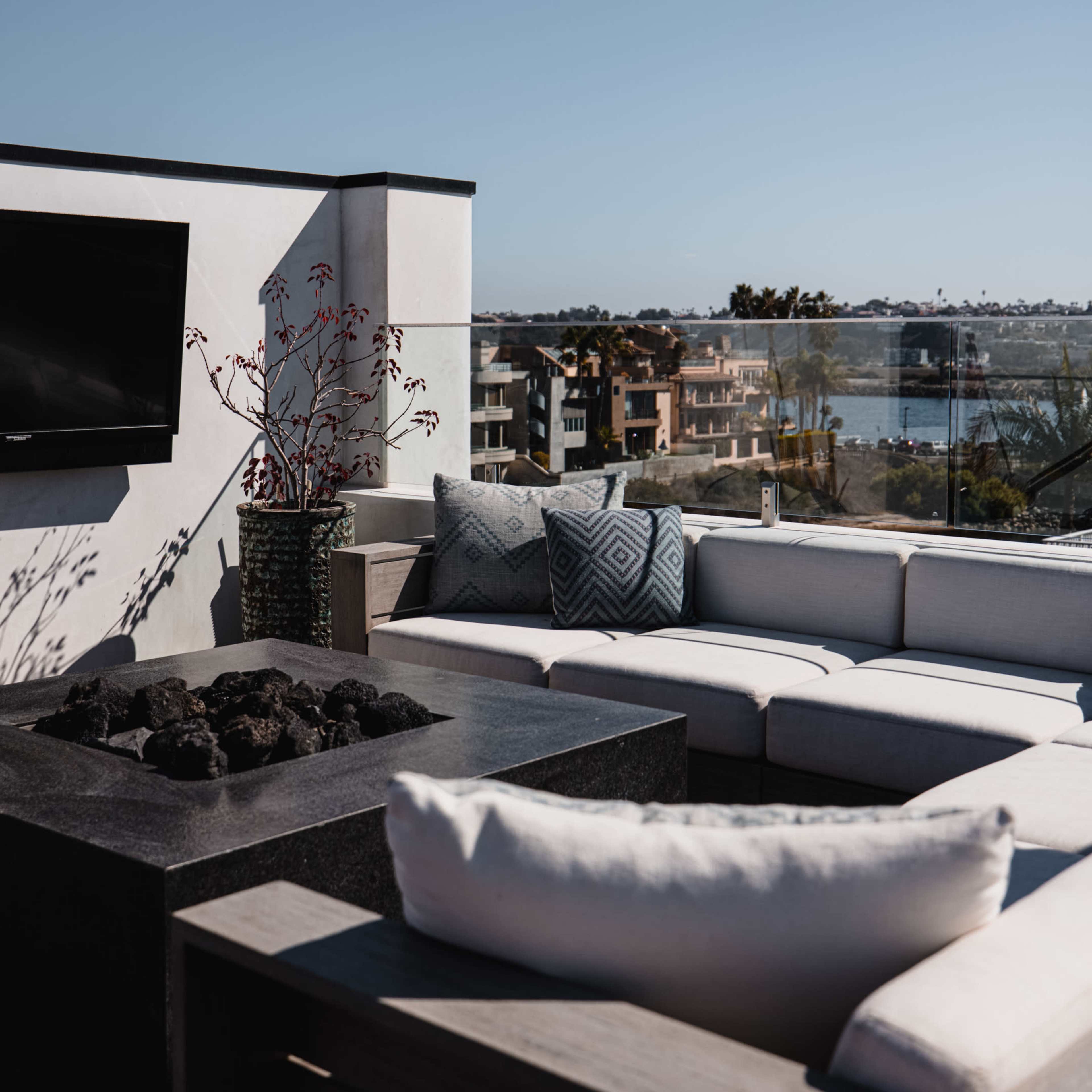 A modern outdoor seating area features a sectional sofa and a fire pit overlooking a body of water and nearby buildings.