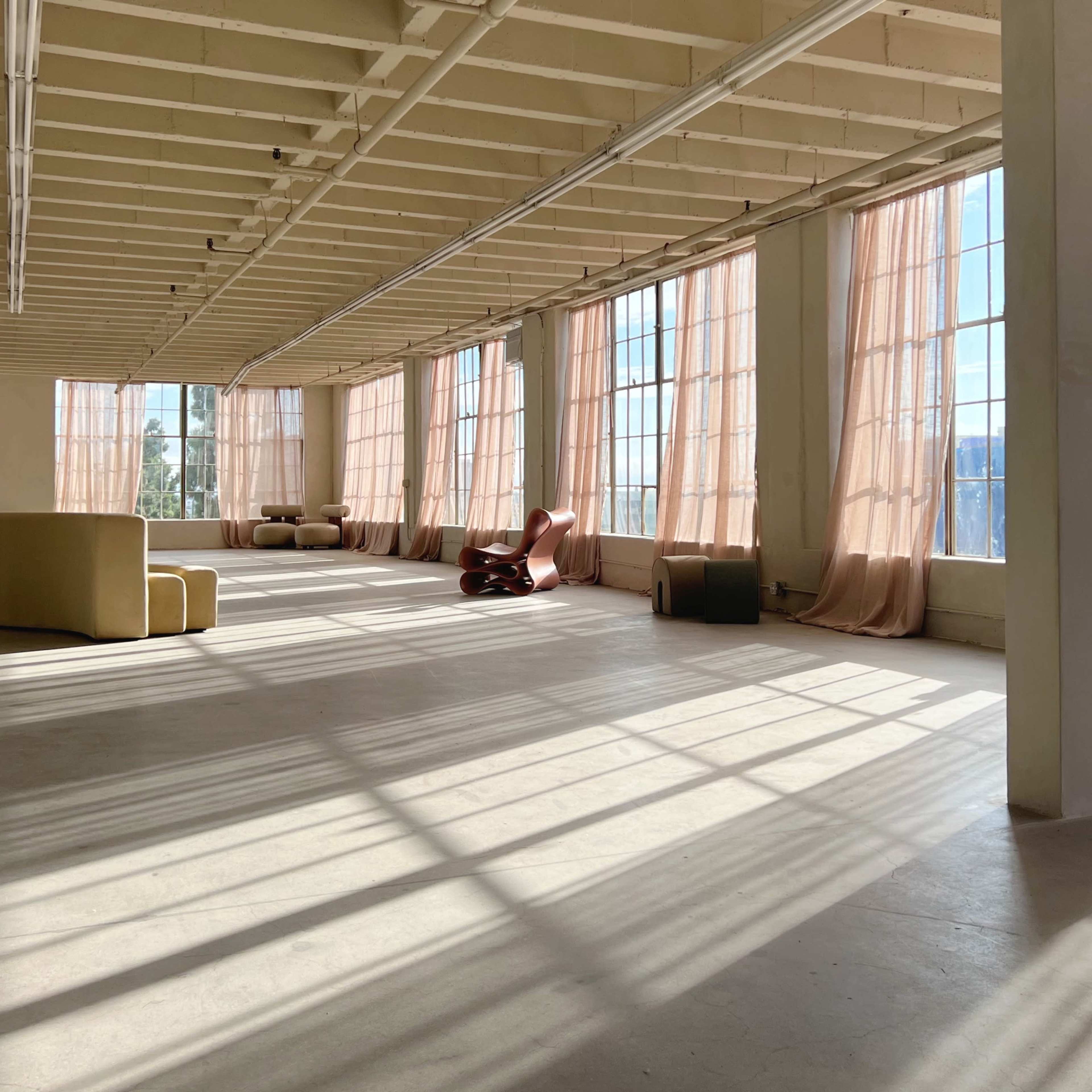 A spacious, sunlit room features large windows with sheer curtains, minimal furniture, and shadow patterns on the floor.