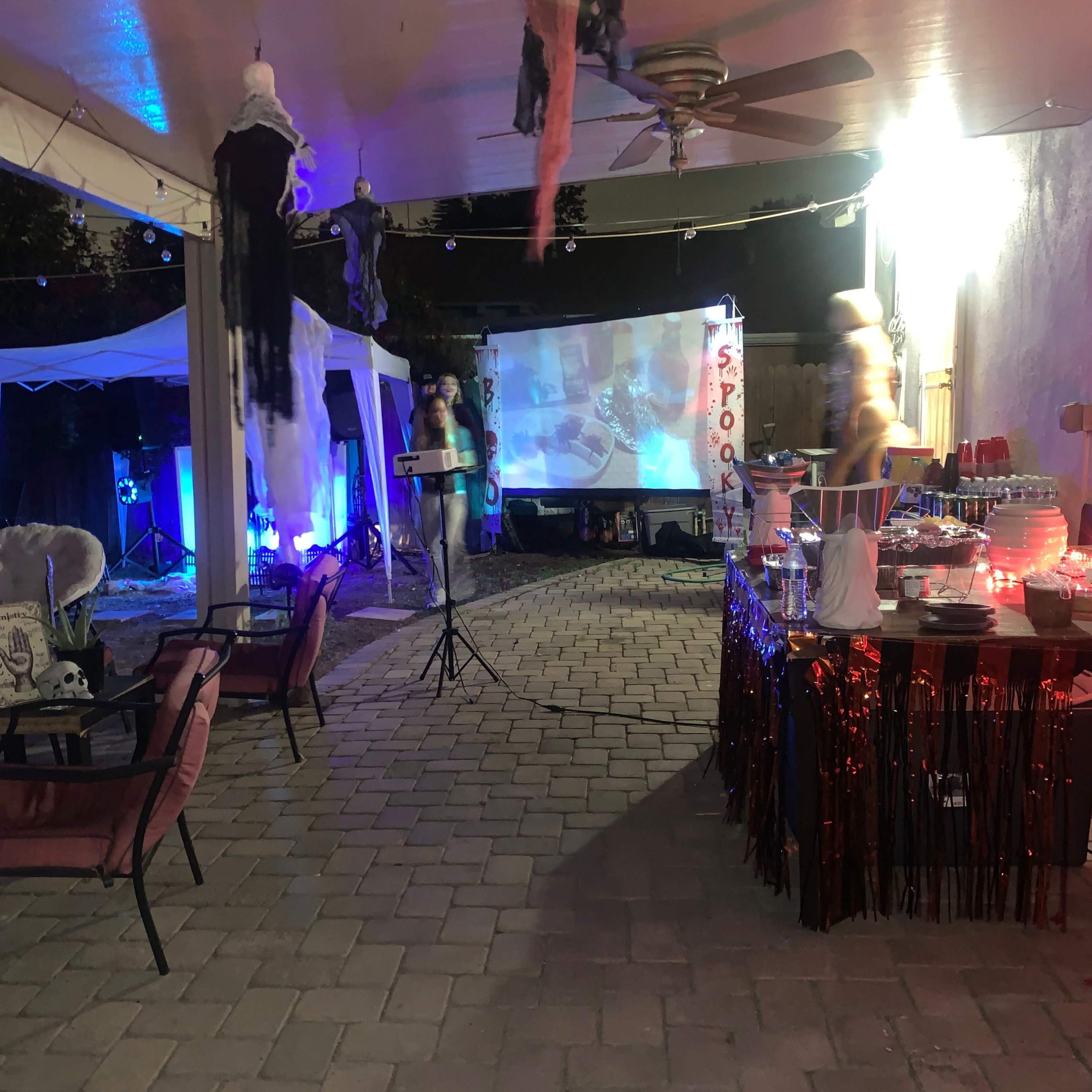 A backyard area is set up for an event, featuring a projector screen, a decorated table, and seating arrangements under a covered patio.