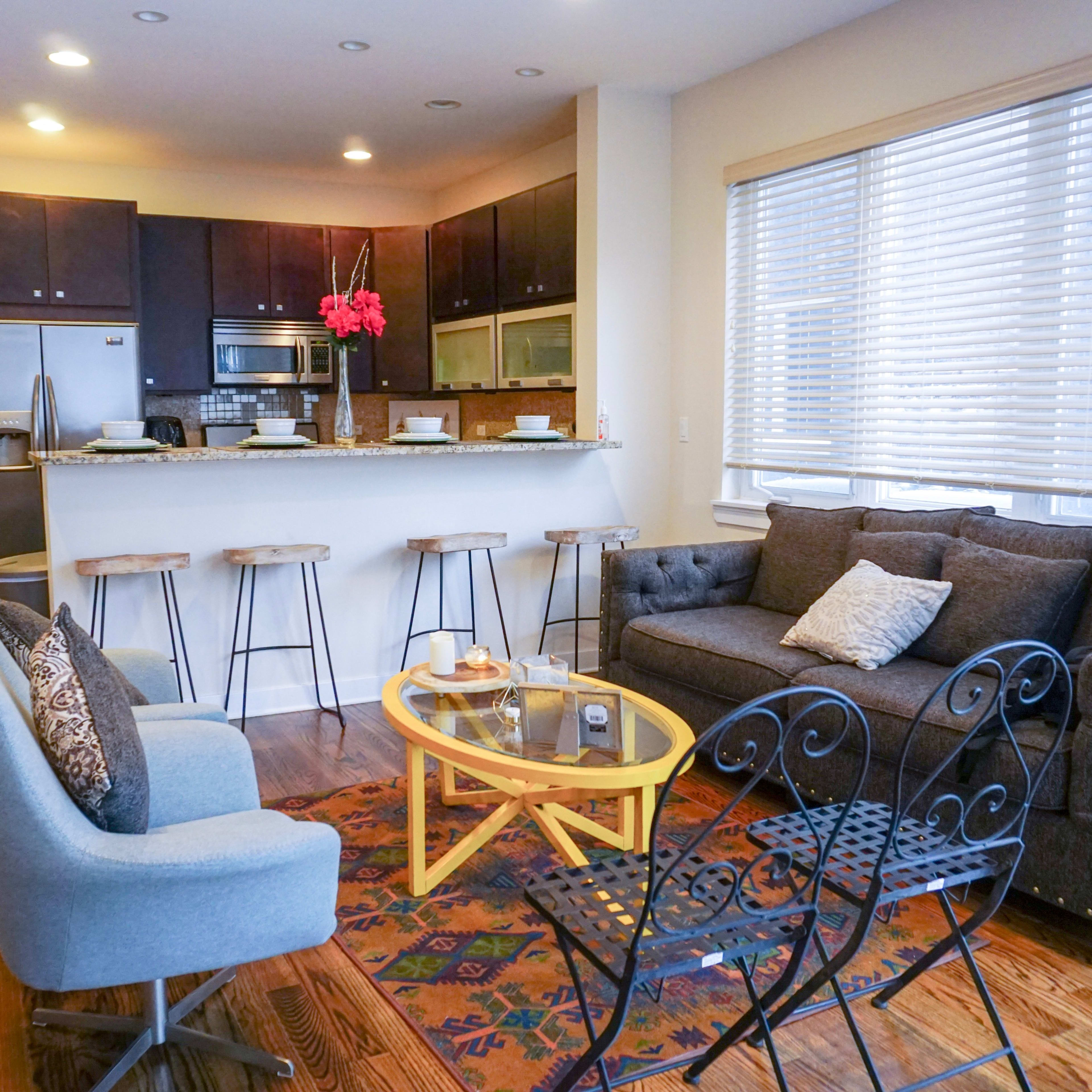 A modern living space features a kitchen bar with stools, a gray sofa, a yellow coffee table, and two outdoor chairs, set against a backdrop of light-colored walls and a hallway.