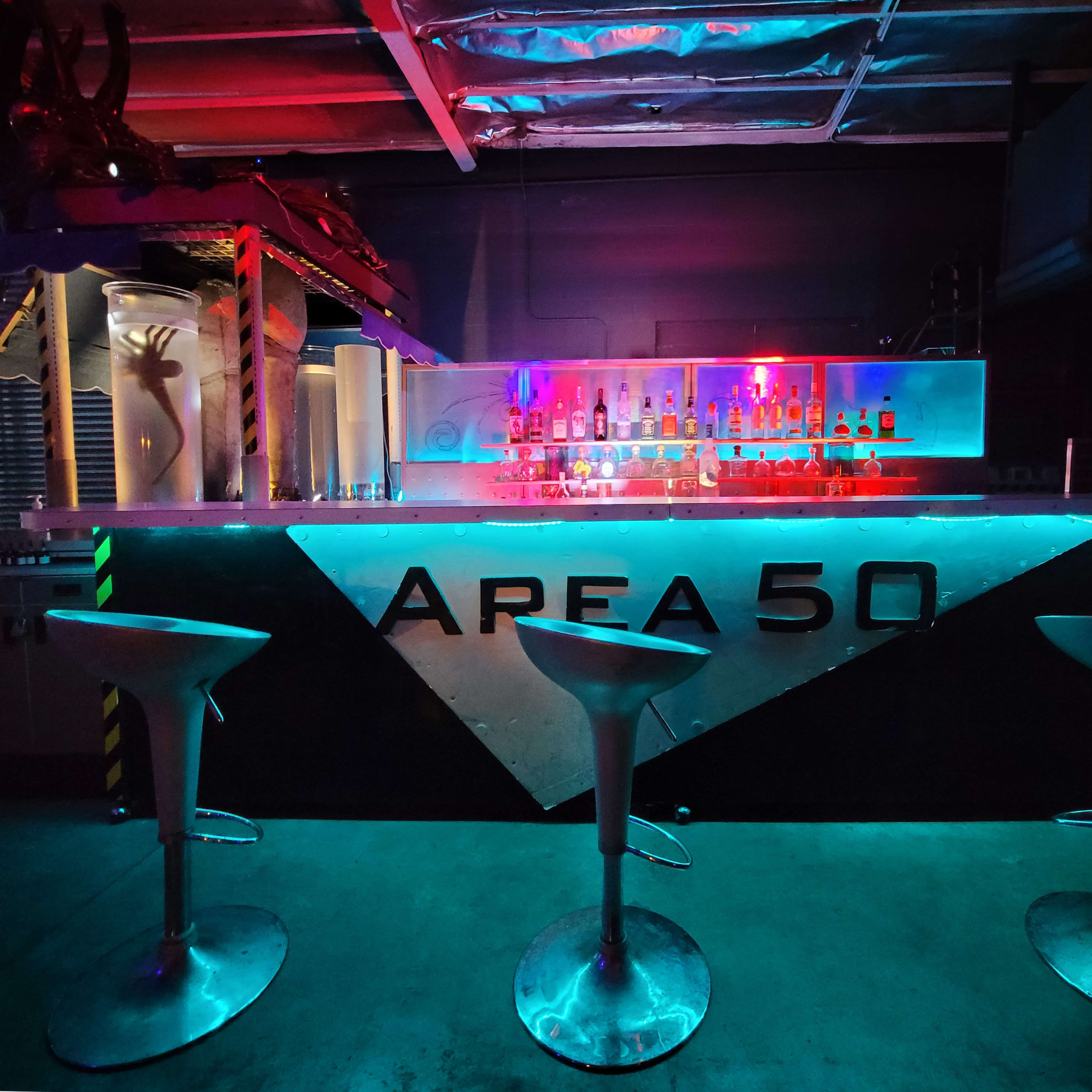 A brightly lit bar with a futuristic design, featuring the name "AREA 50" prominently displayed and accompanied by three metallic stools.