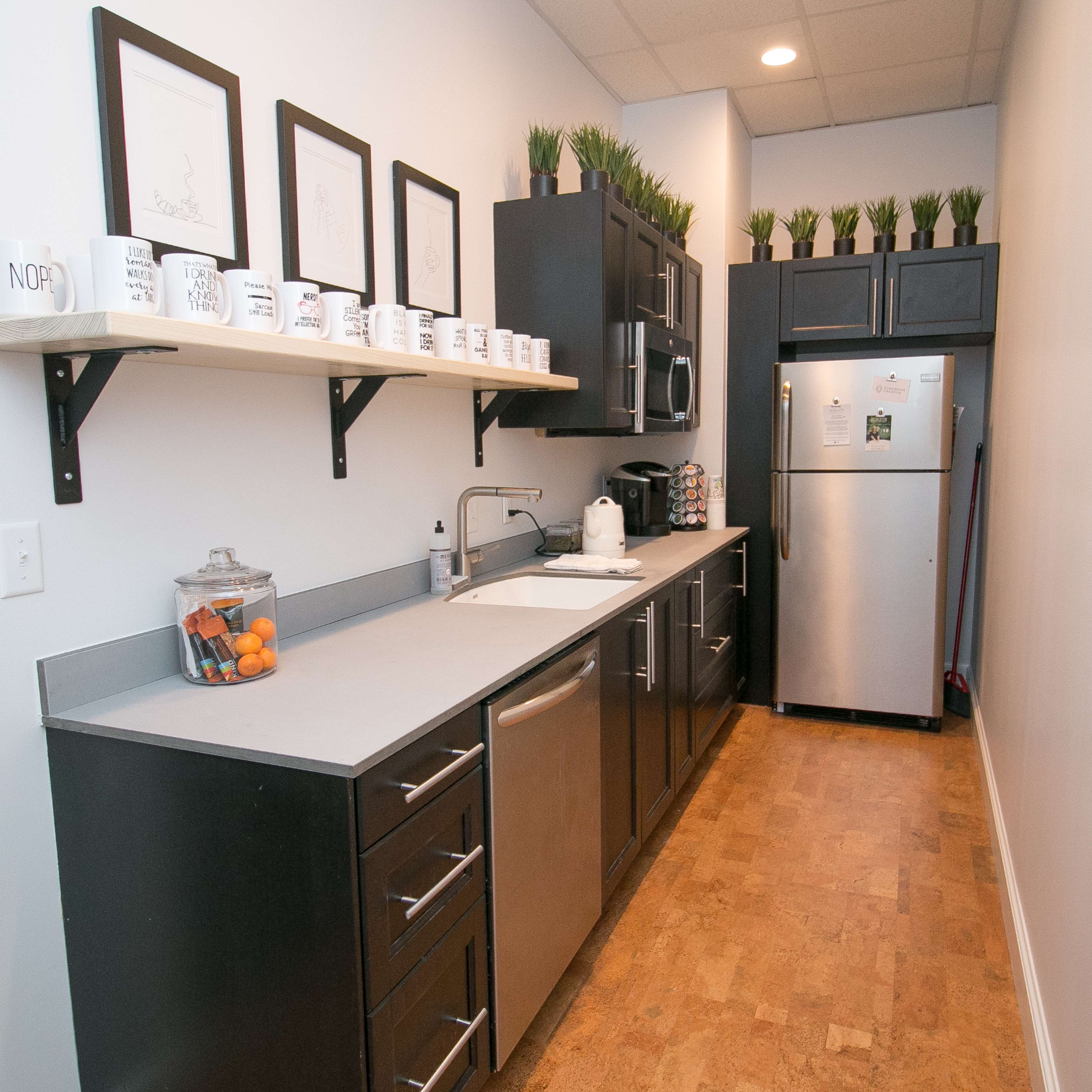 A narrow kitchen with dark cabinetry, a stainless steel refrigerator, and a countertop featuring a sink and a shelf holding mugs and decorative plants.