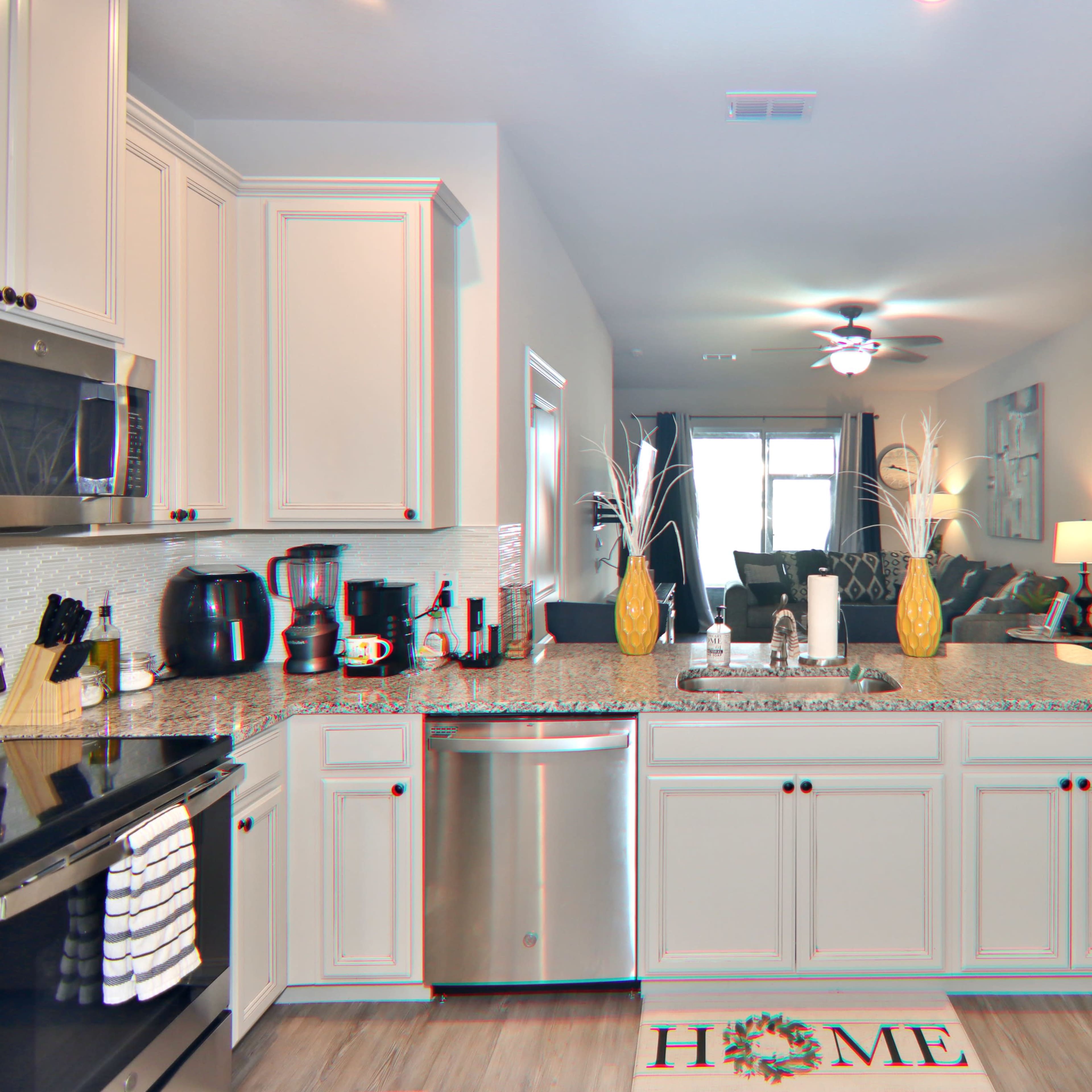 A modern kitchen featuring light-colored cabinets, a stainless steel dishwasher, and a view into a living area with a ceiling fan and dark furnishings.