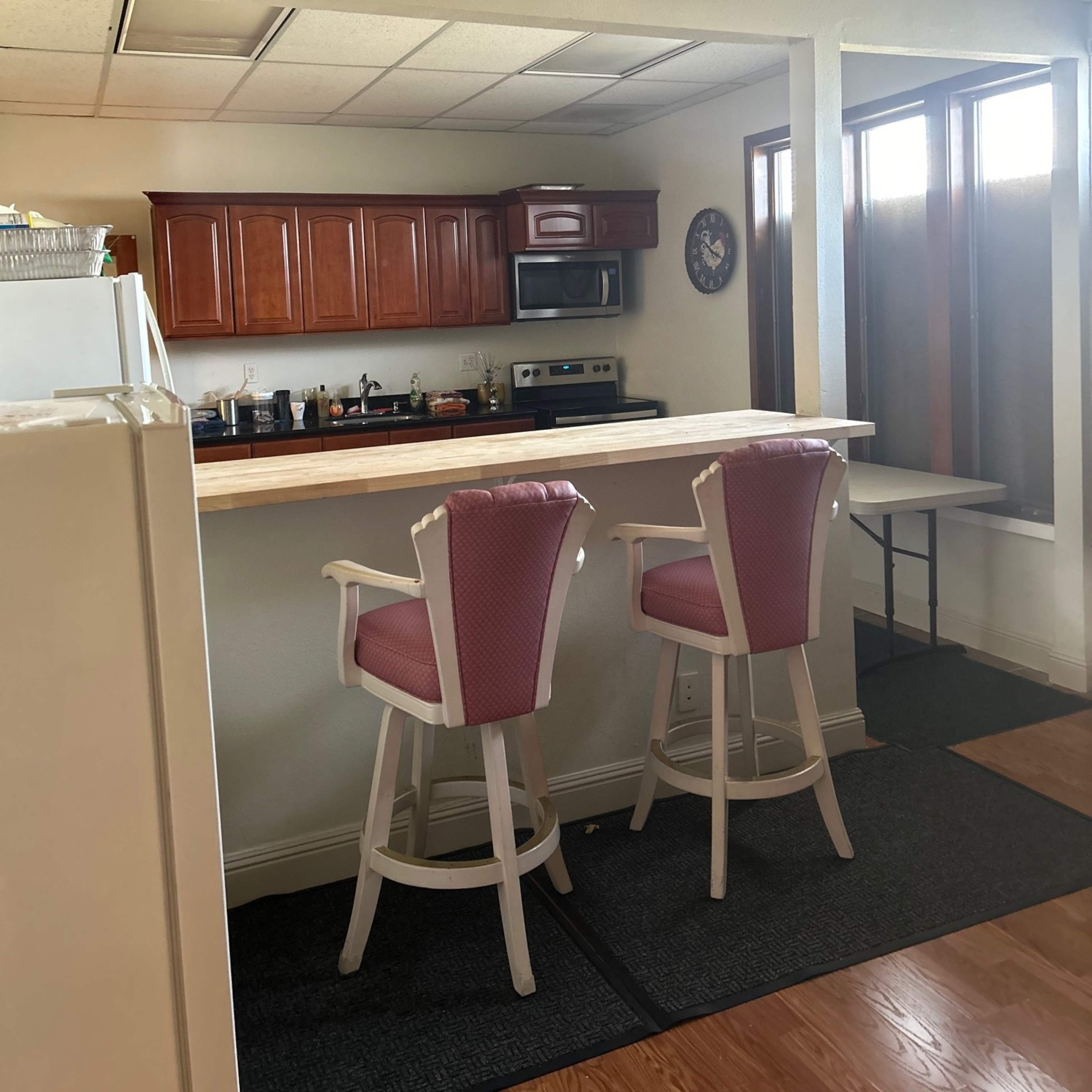 A kitchen area with a bar counter and two pink bar stools, featuring wooden cabinets and a stainless steel microwave in the background.