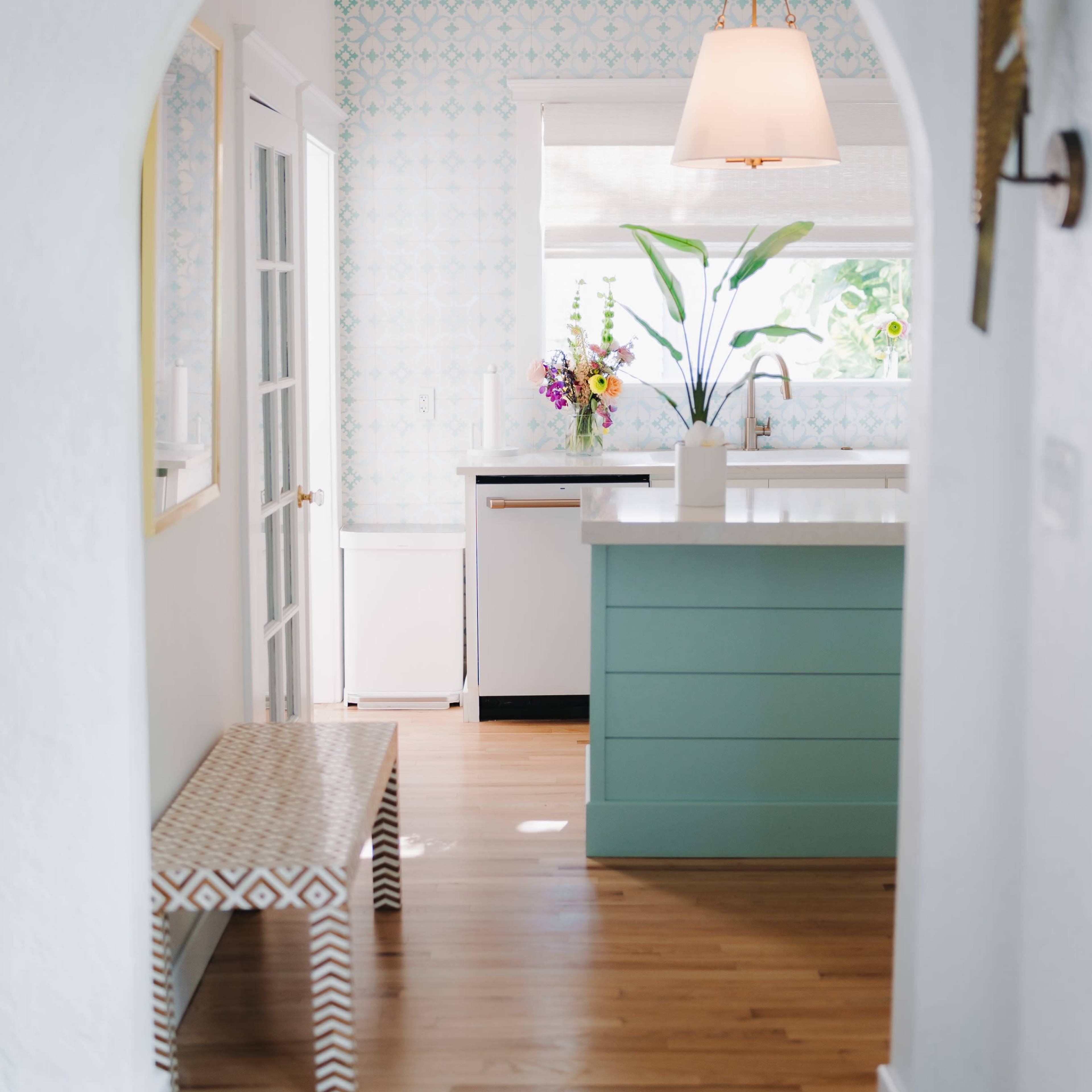 A light-filled kitchen features a pastel blue island, a floral arrangement on the counter, and an archway leading to a hallway.