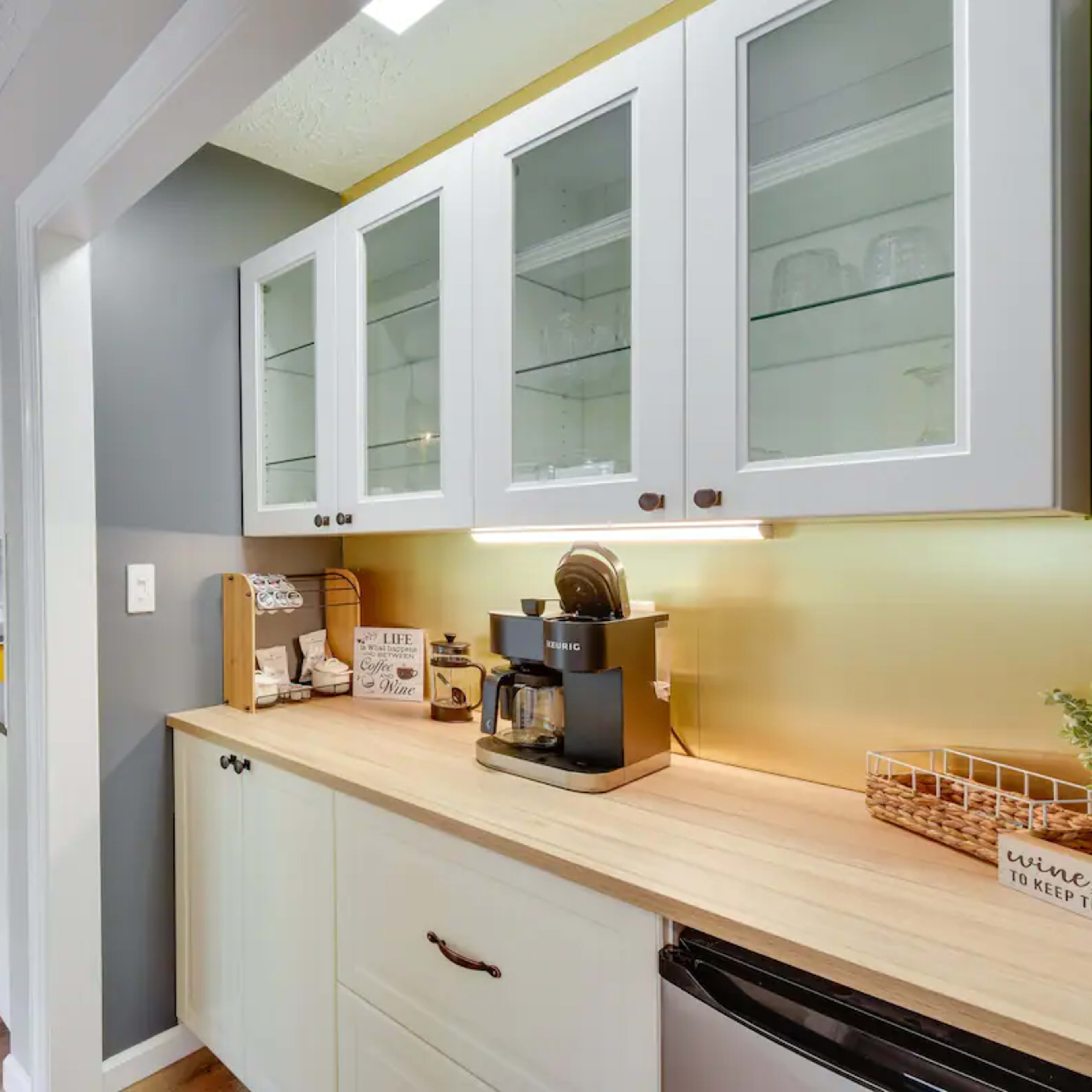 A modern kitchen area featuring white cabinets with glass doors, a wooden countertop with a coffee maker, and a bright yellow cabinet in the background.