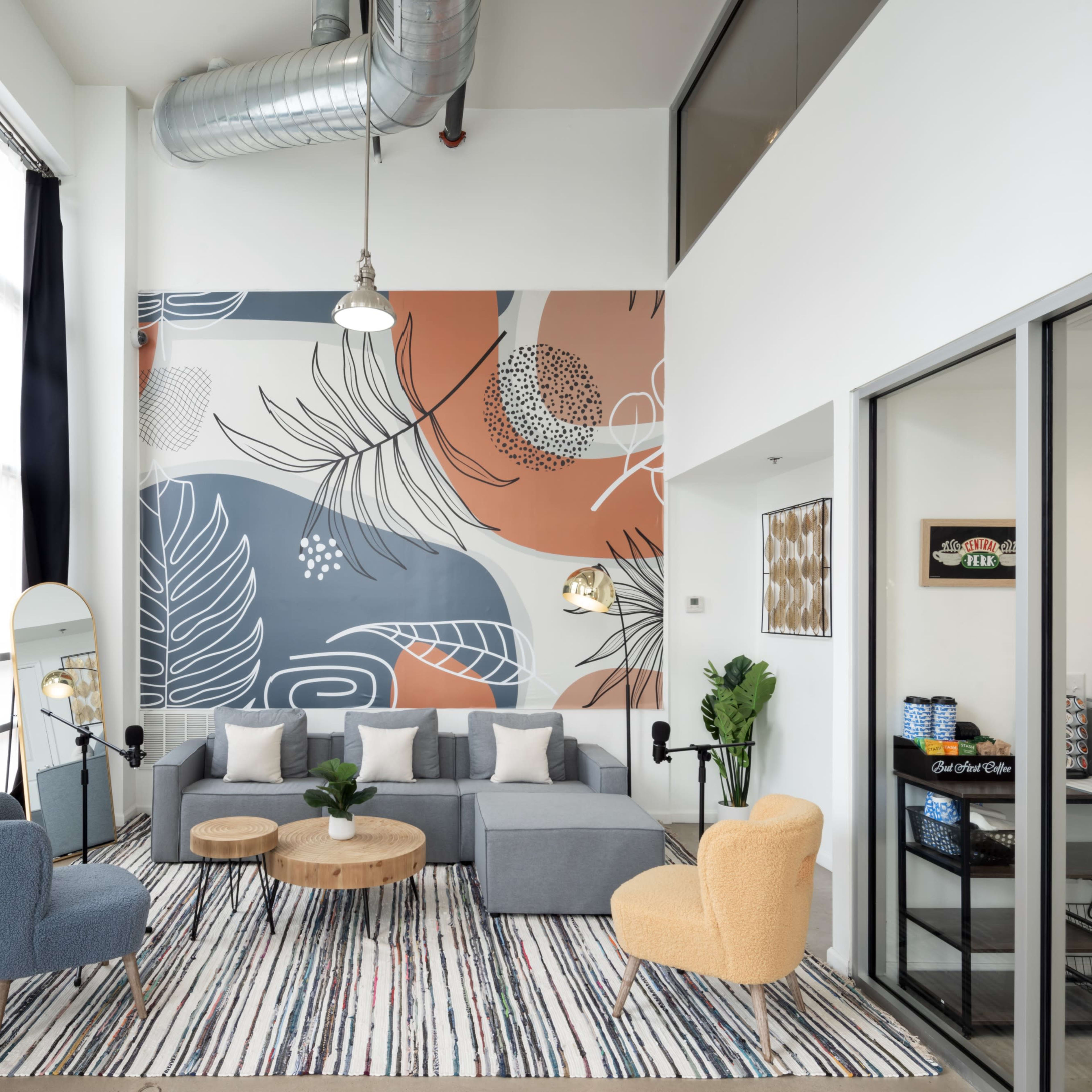 A modern living room decorated with a patterned accent wall, a gray sectional sofa, two colorful armchairs, a round wooden coffee table, and various indoor plants.
