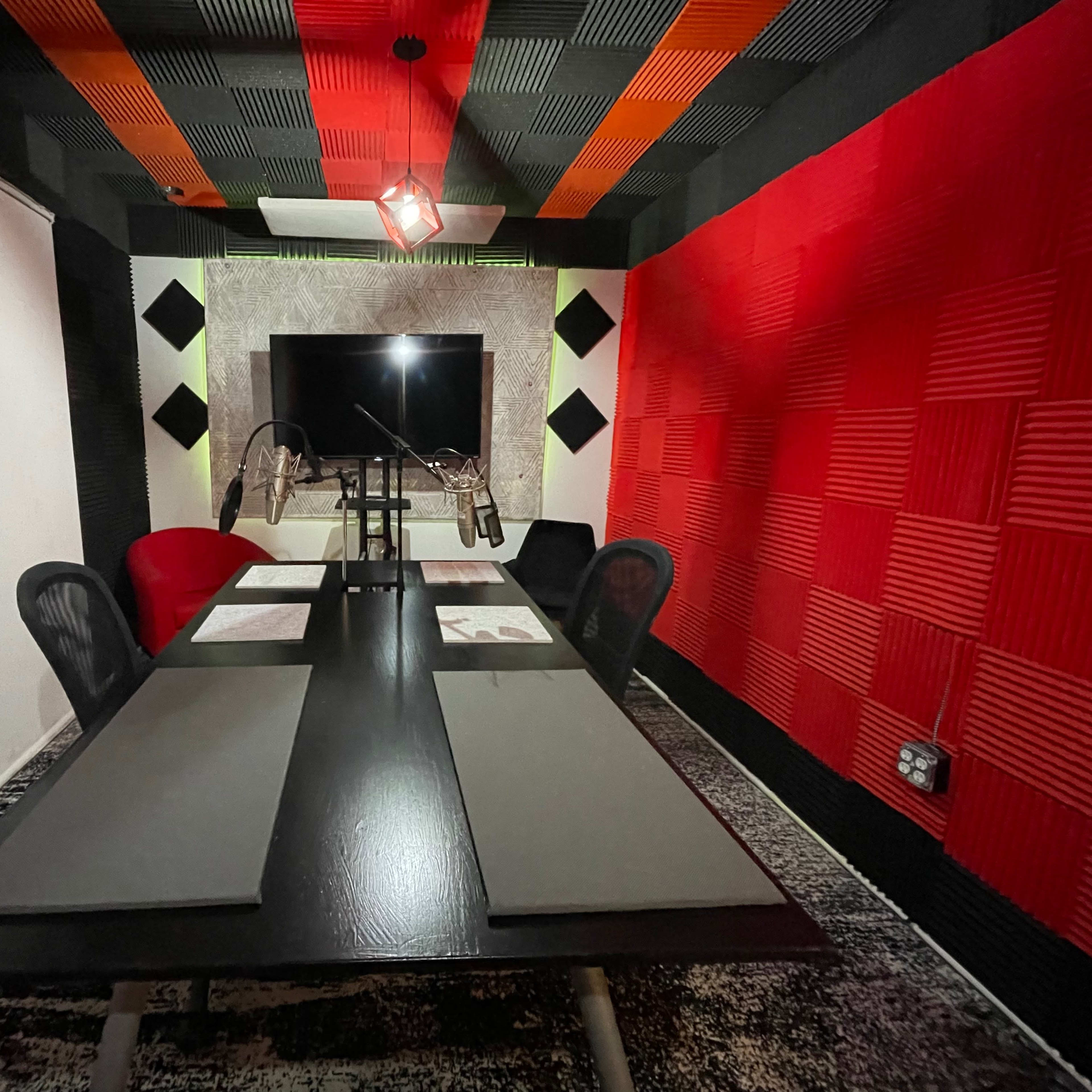A soundproof recording studio features a long black table with chairs, red and black acoustic foam on the walls, and a large screen at the far end.