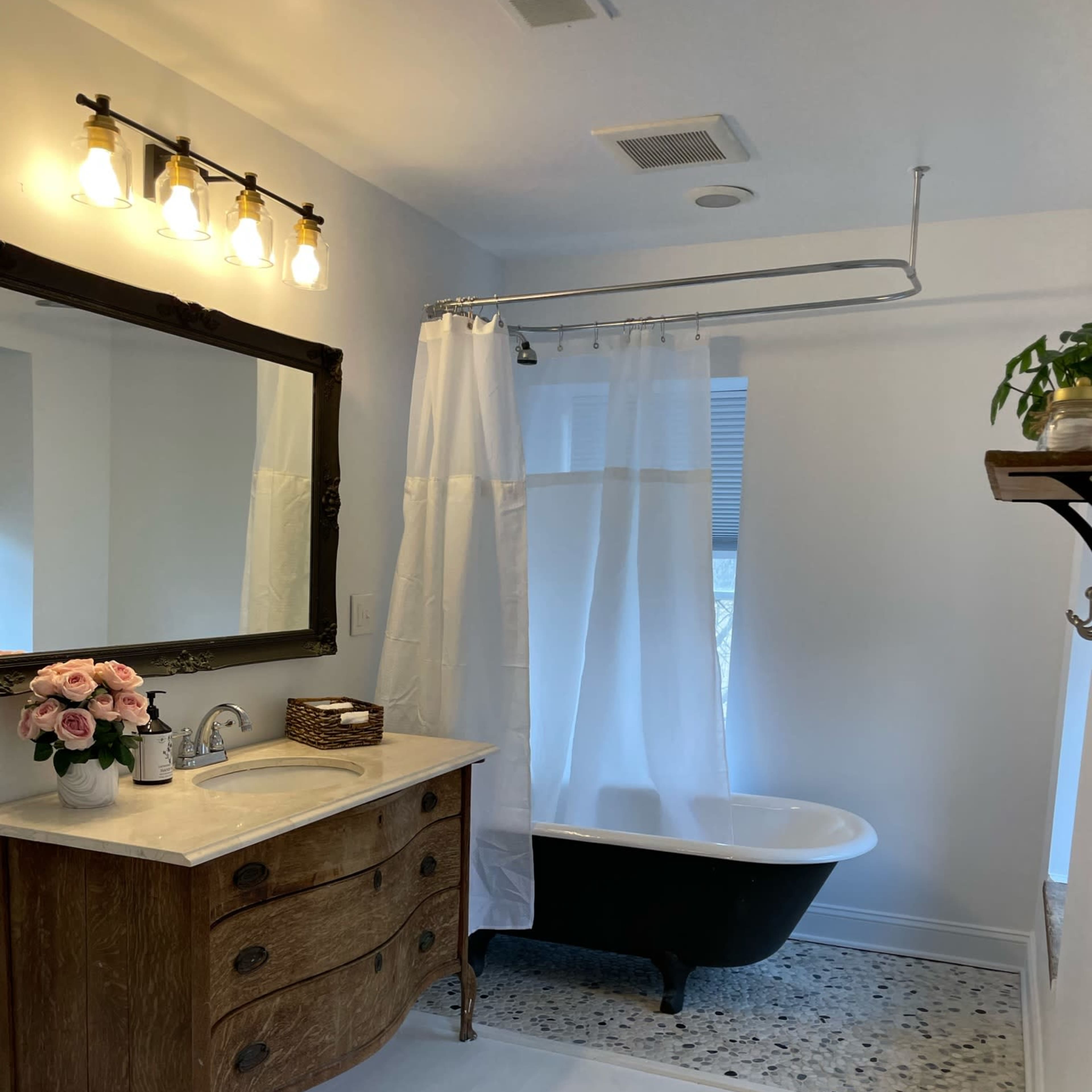 The bathroom features a clawfoot tub, a large mirror above a wooden vanity, and a white shower curtain, complemented by a floral arrangement and pebble flooring.
